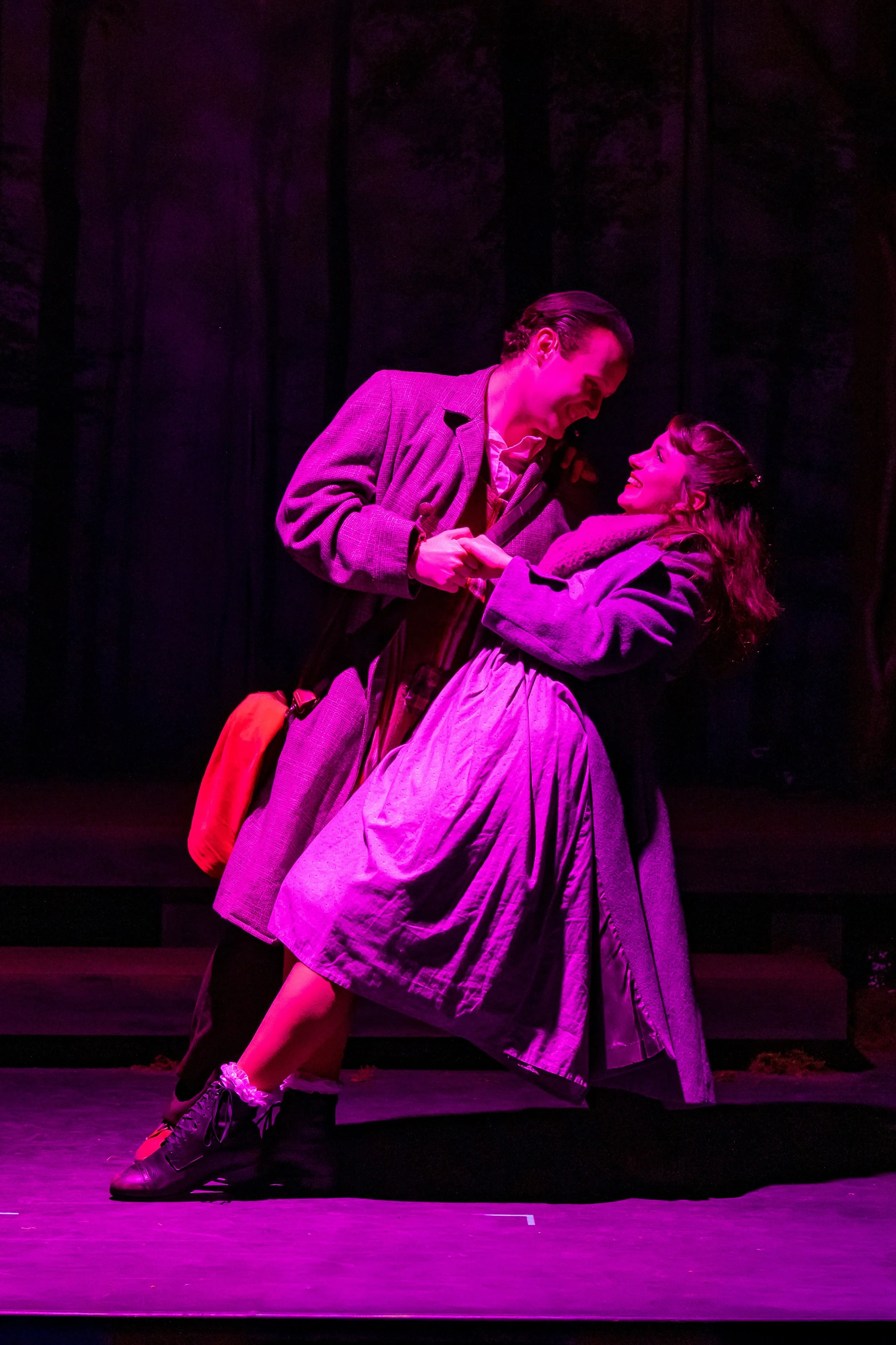 Two actors performing a scene on stage, lit with pink and purple lighting, dressed in period costumes, appears to be a theatrical play.