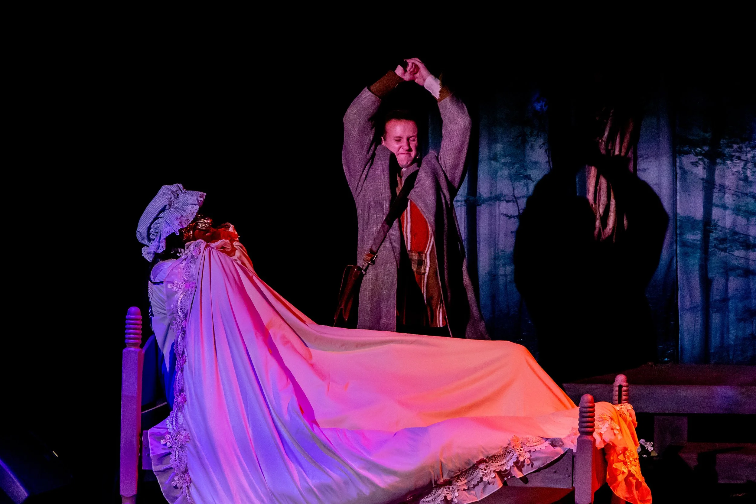 Actors performing on stage, one man with hands raised and two women in period costumes, with a forest backdrop.
