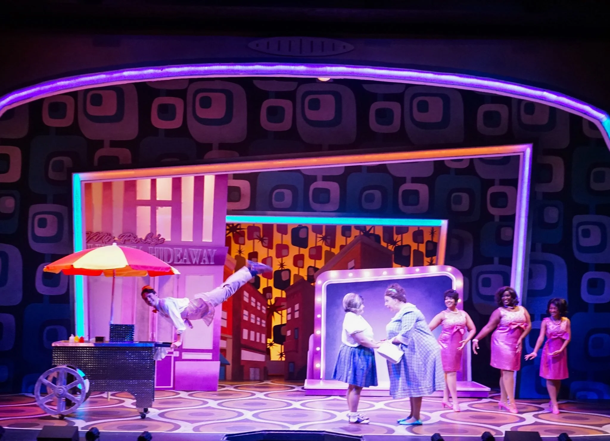 Stage scene from the musical 'Nice Pink', featuring five performers and a vibrant backdrop with cityscape and colorful patterns, including a man mid-air performing an acrobatic move.