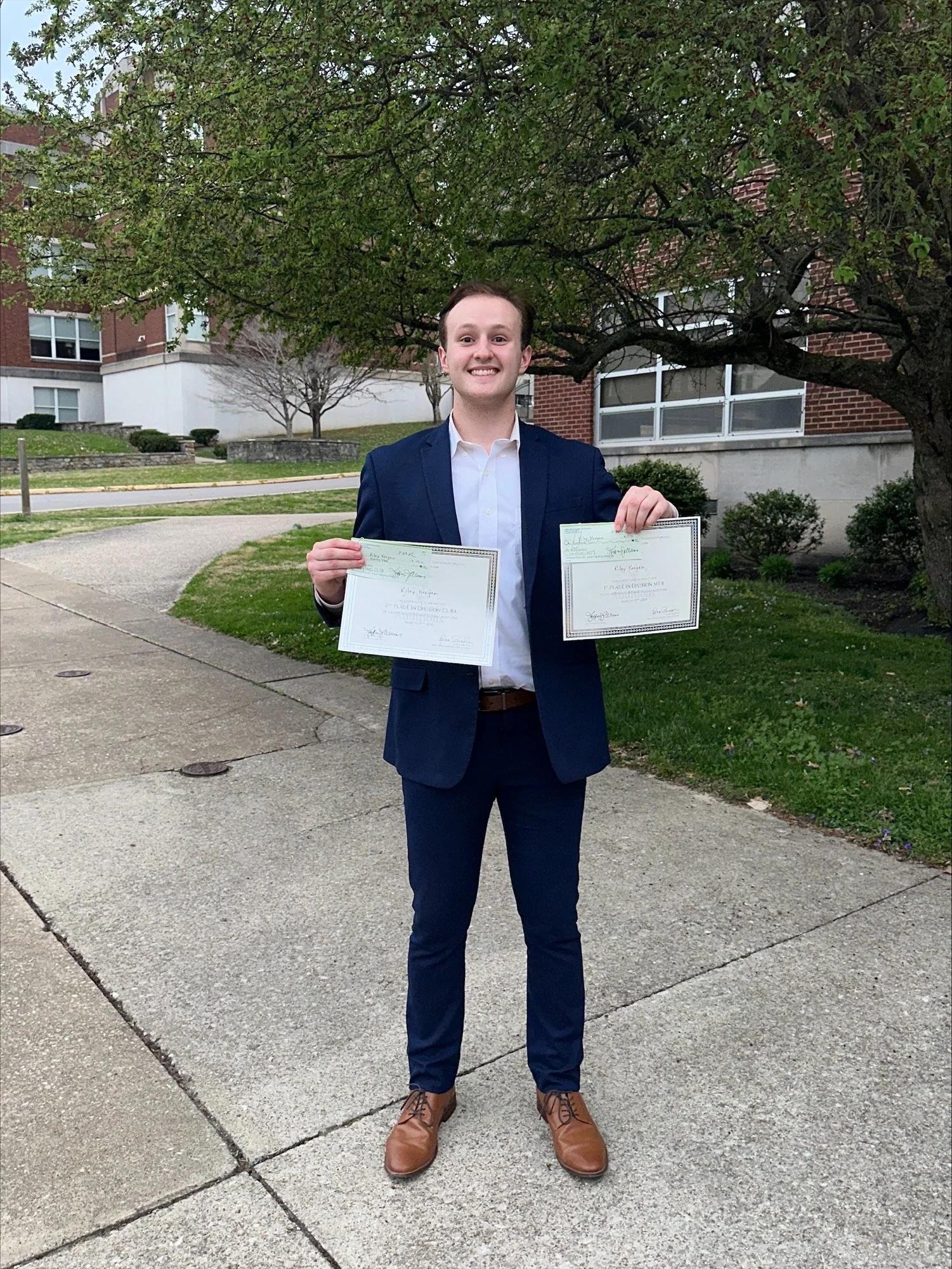 Young man in navy suit holding diplomas outside near a tree, smiling.