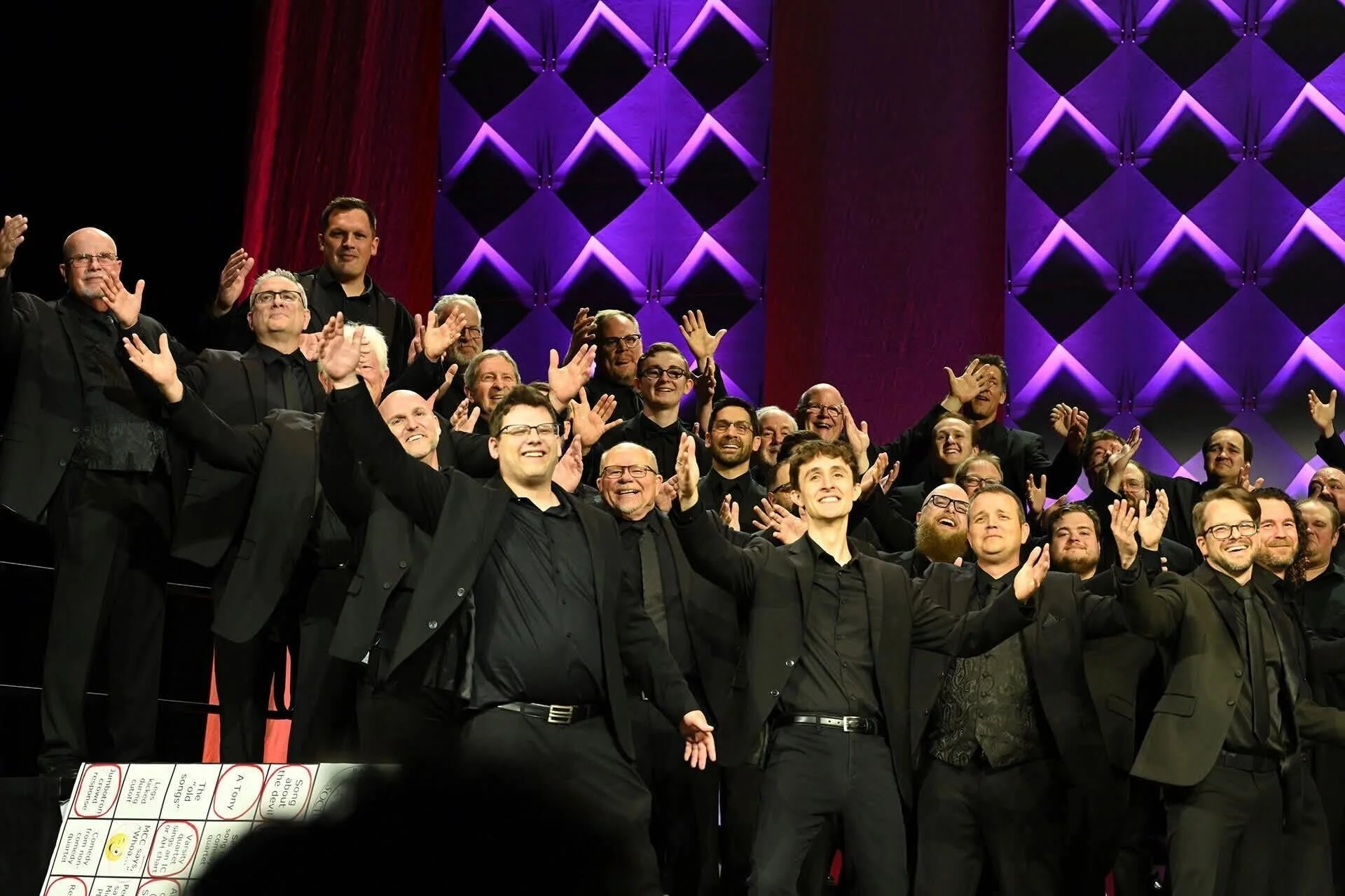 Photograph of a large choir or group of singers dressed in black suits, standing on stage with purple lit backdrop, smiling and raising their hands in celebration.