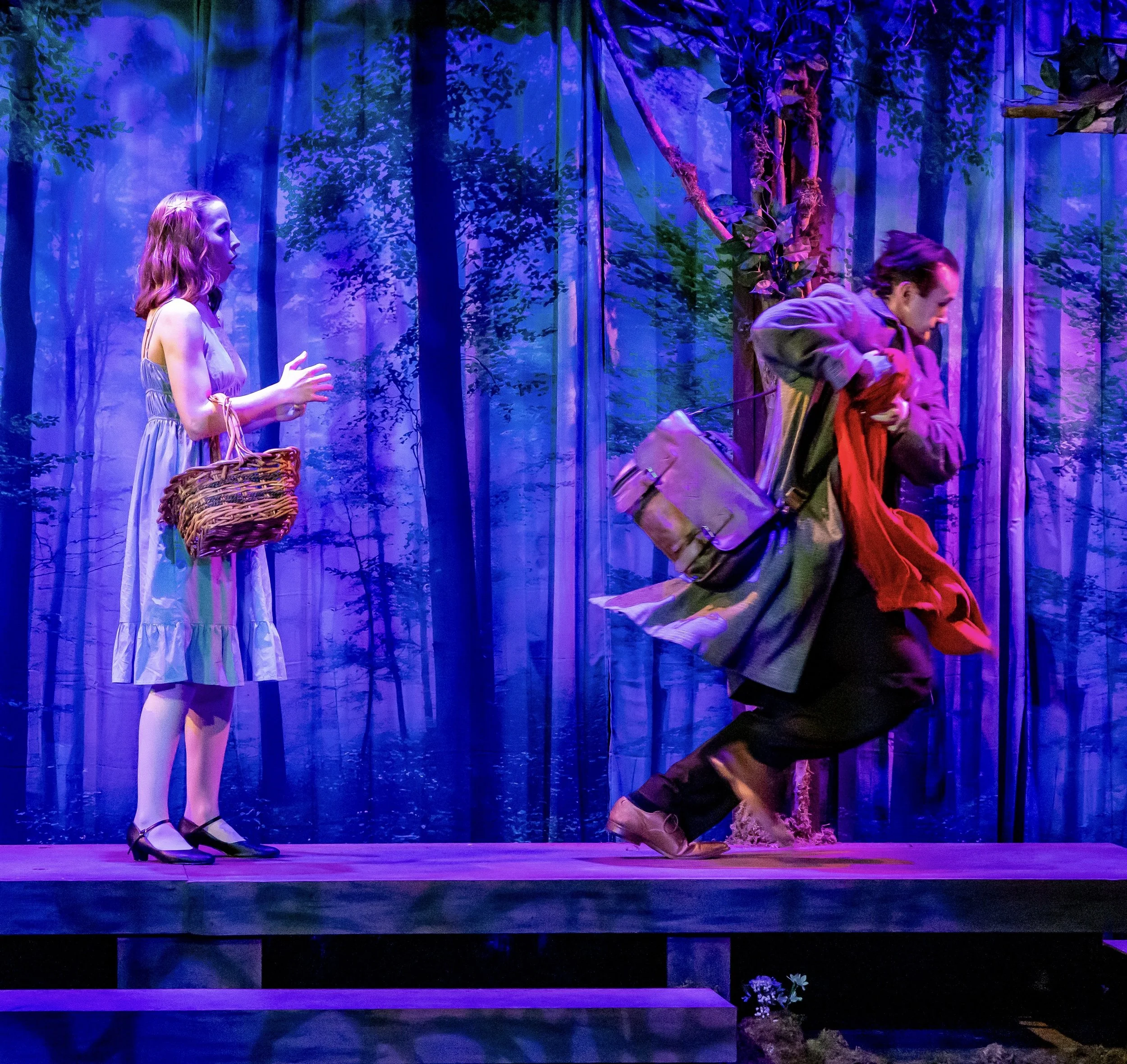 Two actors on stage, one woman with a basket talking to a man running away in a forest scene, with purple and blue forest backdrop.