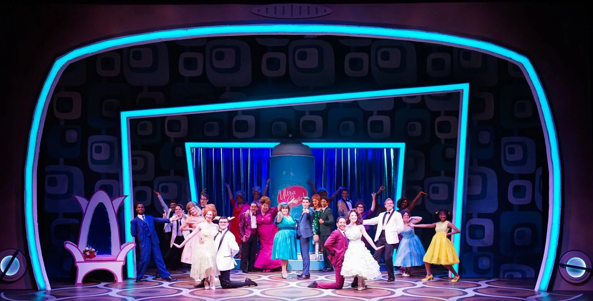 Stage scene from a musical with a colorful set, featuring a diverse cast of performers in period costumes, standing in lively poses.