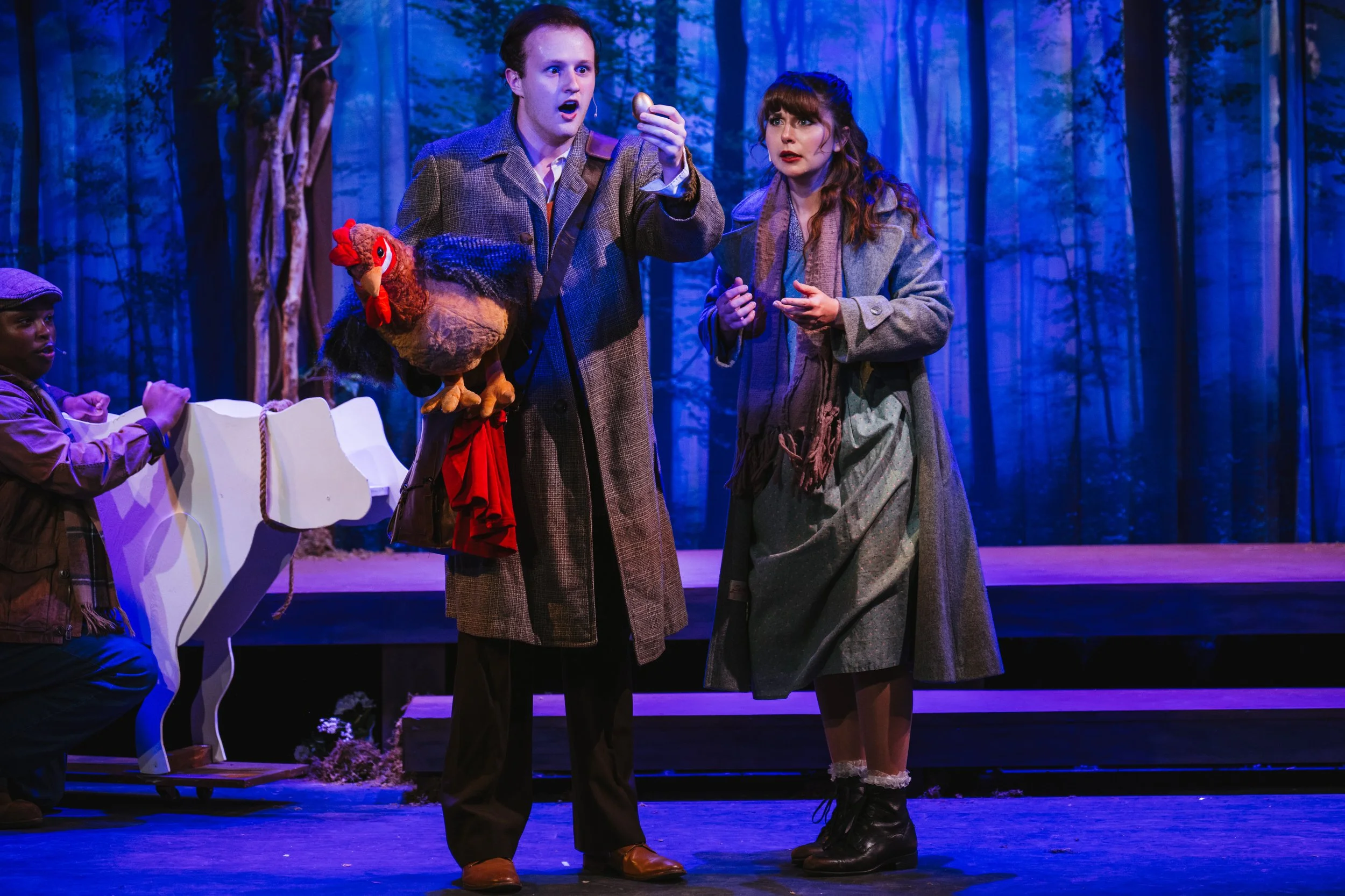 Two actors in period costumes perform on stage in a theatrical production set in a forest, with one holding a puppet of a chicken.