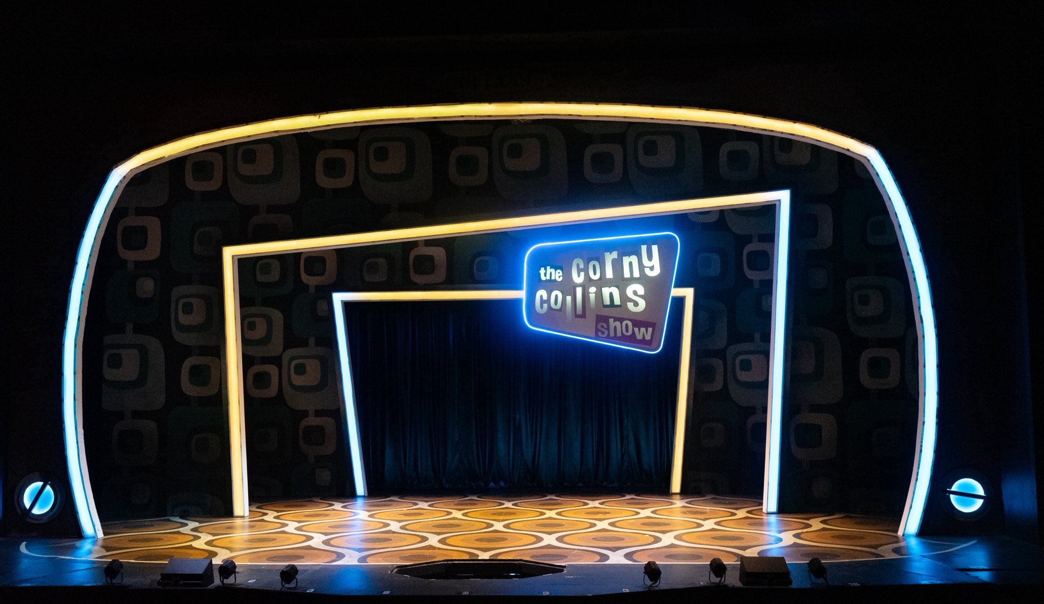 A stage with a neon sign that reads 'the corny collins show' and a patterned floor, set for a performance, framed by colorful neon lights.