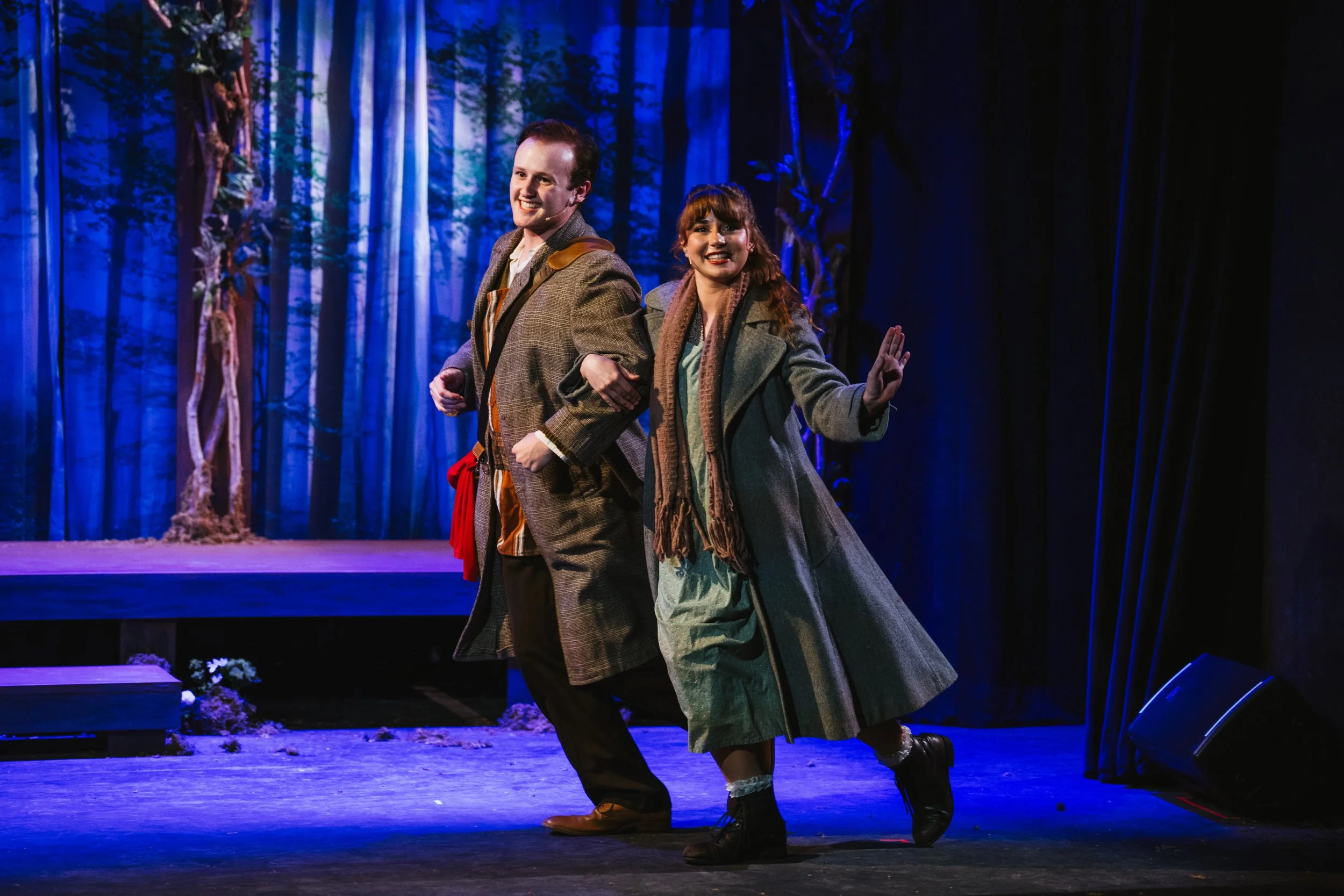 Two actors on stage performing, dressed in period costumes, standing in front of a blue forest background, with one actor smiling and the other waving.