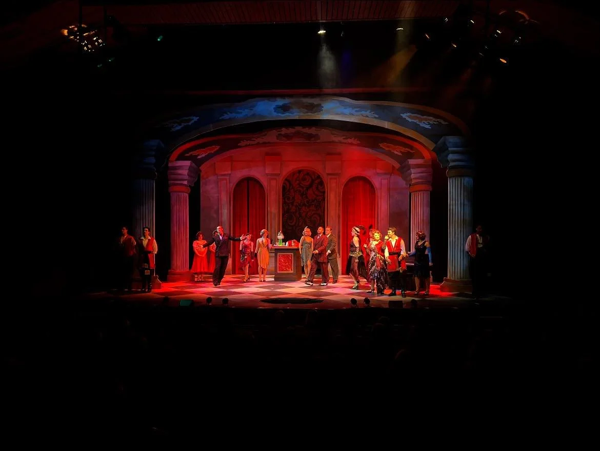 A theatrical stage with actors performing a scene. The set resembles a grand room with red walls, columns, and decorative elements. The actors are dressed in vintage or formal costumes, some holding drinks, suggesting a lively social gathering.
