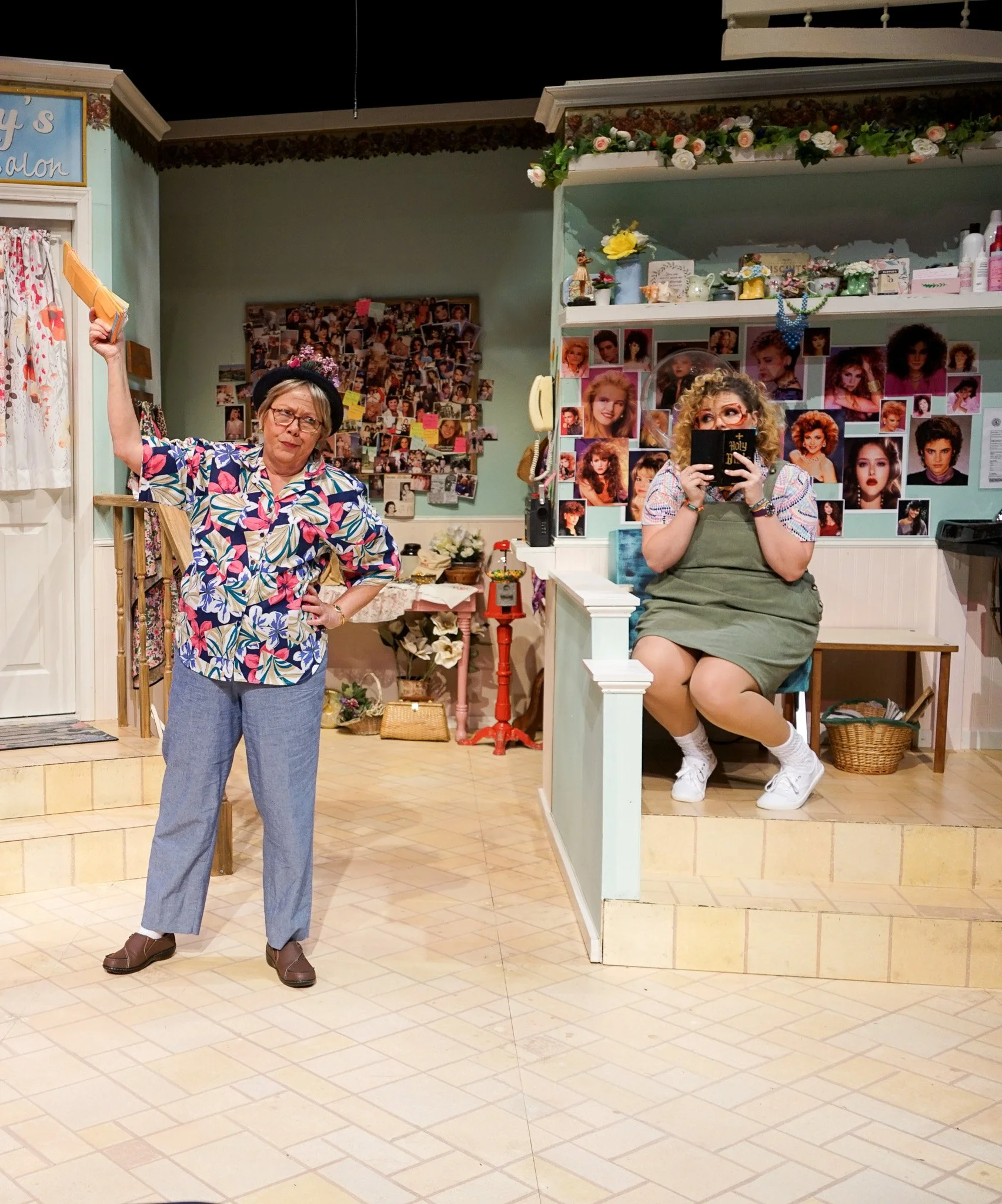 Stage scene from a play or musical with two women in a hair salon set. One woman stands on the left holding a yellow item, wearing a colorful floral blouse, jeans, and a hat, with a puzzled expression. The other woman sits on the right in a green apr