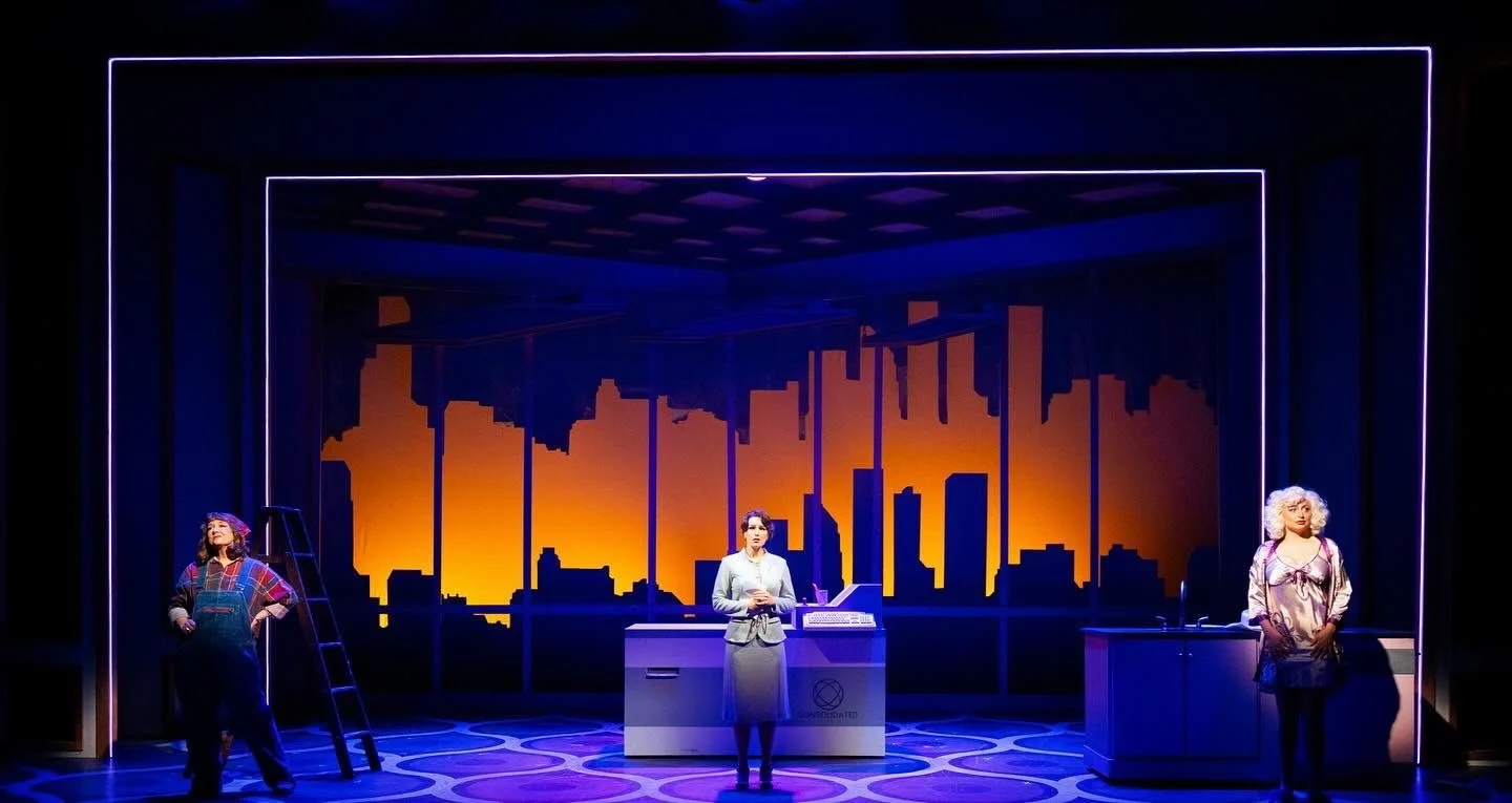 Stage scene with three women standing in front of a city skyline backdrop at sunset, illuminated by neon lights.