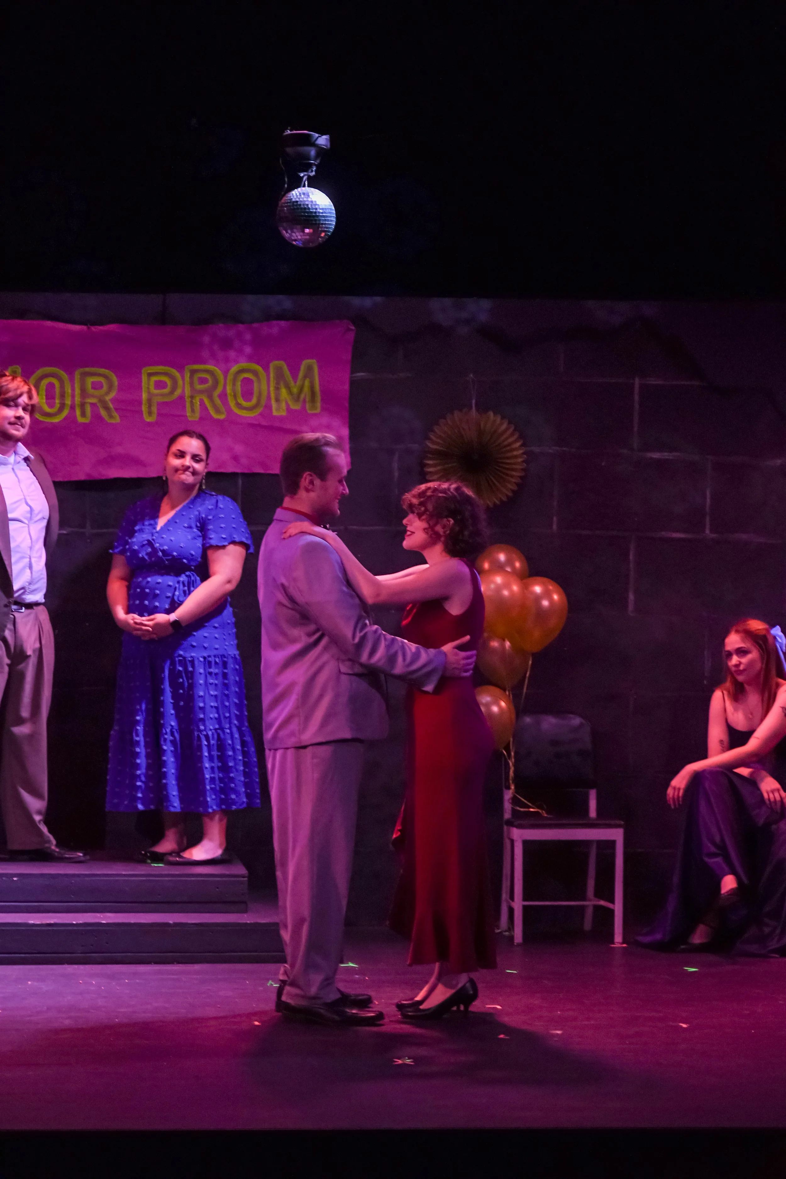 Scene from a prom with people on stage dancing and talking, decorated with balloons, a banner, and a disco ball hanging from the ceiling.