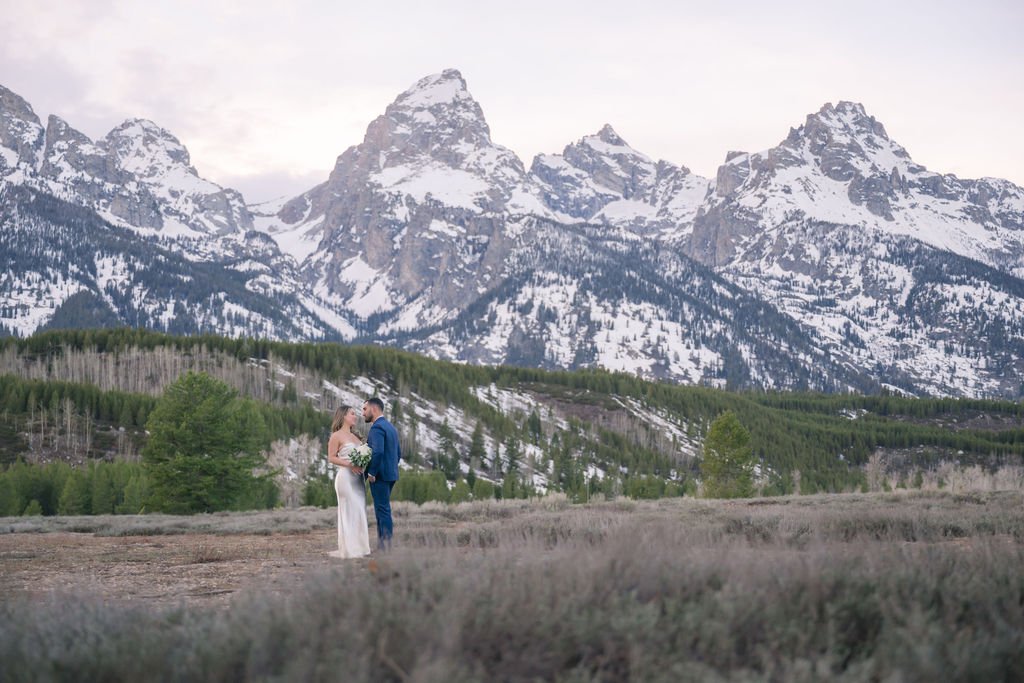 bride and groom in front of the grand tetons.  Bride is holding a bouquet in a white dress and groom is wearing a blue tux