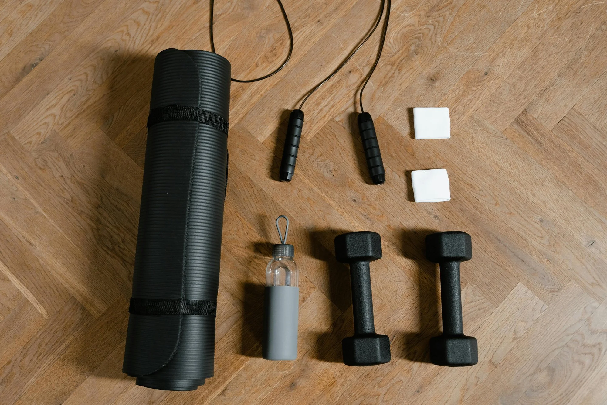 Home gym workout equipment including a rolled yoga mat, jump rope, two foam pads, a water bottle with a silicone sleeve, and two dumbbells on a wooden floor.