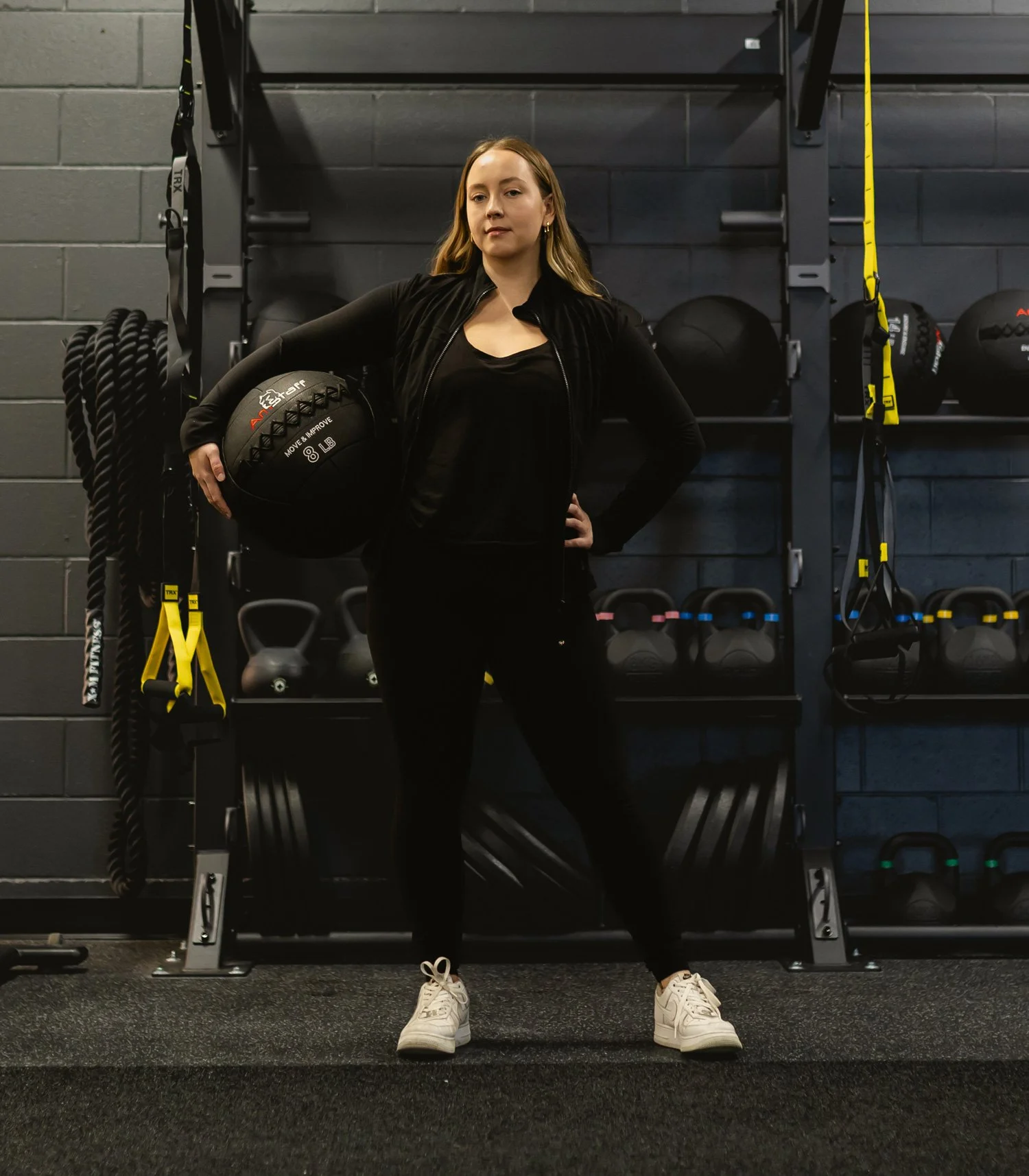 A woman standing in a gym, holding a black medicine ball with her left arm, with fitness equipment and black wall behind her. EverActive Boutique Fitness Walkerton, Physiotherapist Led Exercise, Functional Strength Training, Post-Rehab Wellness Ont