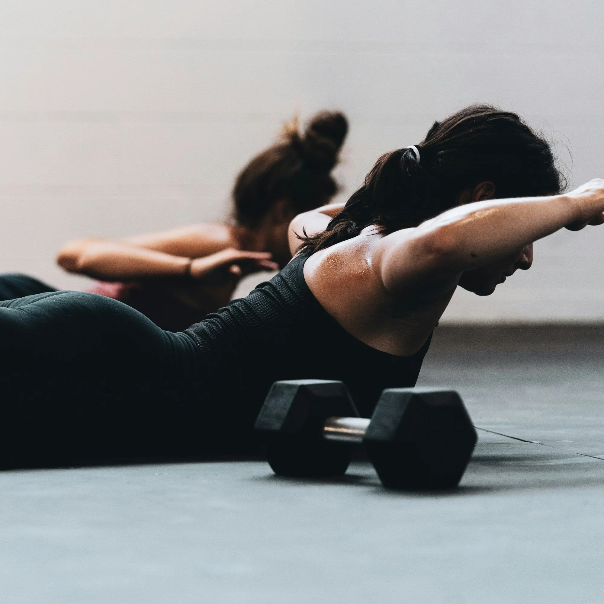 Two women exercising on the floor with dumbbells nearby, doing a plank position with arms extended.
