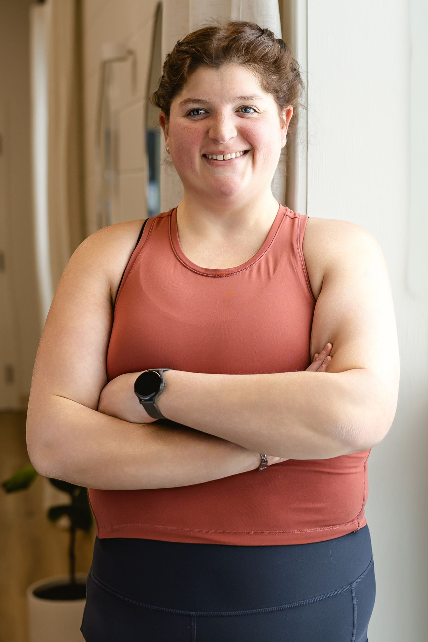 A woman with brown hair, wearing a sleeveless reddish top and black leggings, standing indoors with her arms crossed, smiling near a window with white curtains. EverActive Walkerton, Physiotherapist Led Exercise.