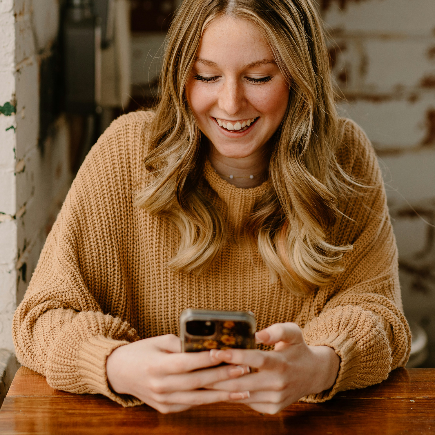 A young woman with long, wavy blonde hair and a big smile, wearing a tan sweater, looking at her smartphone while sitting at a wooden table.