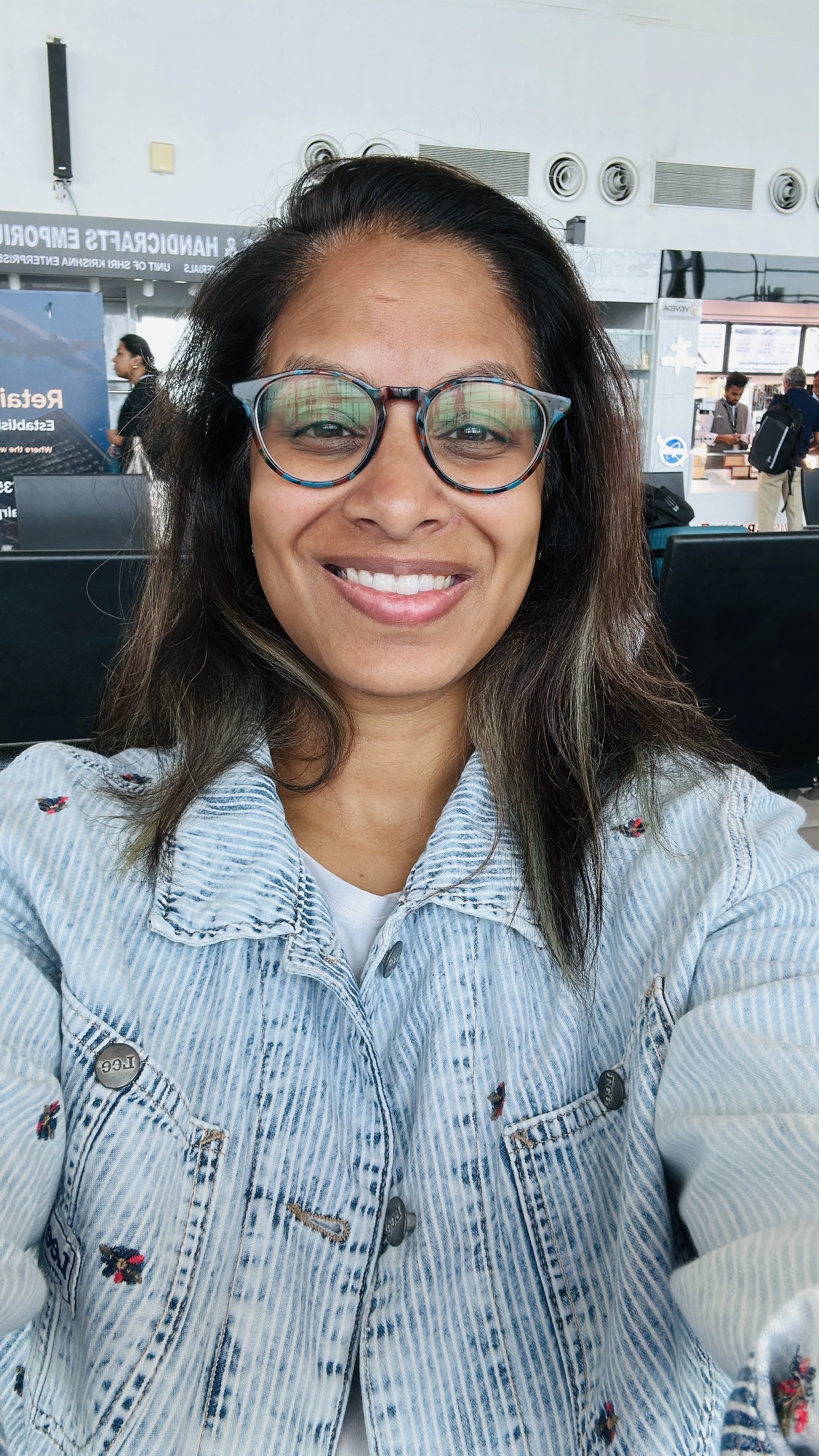 Thought leader and parent, Ponni Carlin, is smiling while sitting in an airport or transportation hub, wearing glasses and a light blue striped denim jacket.