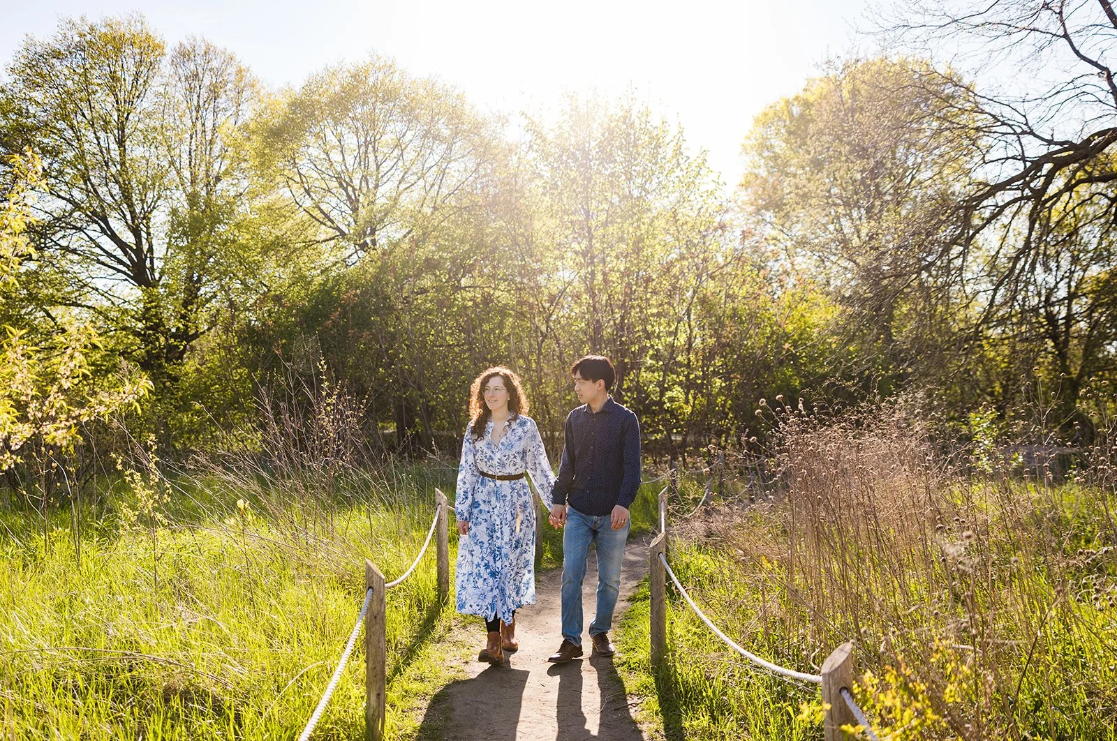 A young couple walking hand-in-hand on a narrow dirt path in a sunlit park or garden with trees and tall grass on either side.