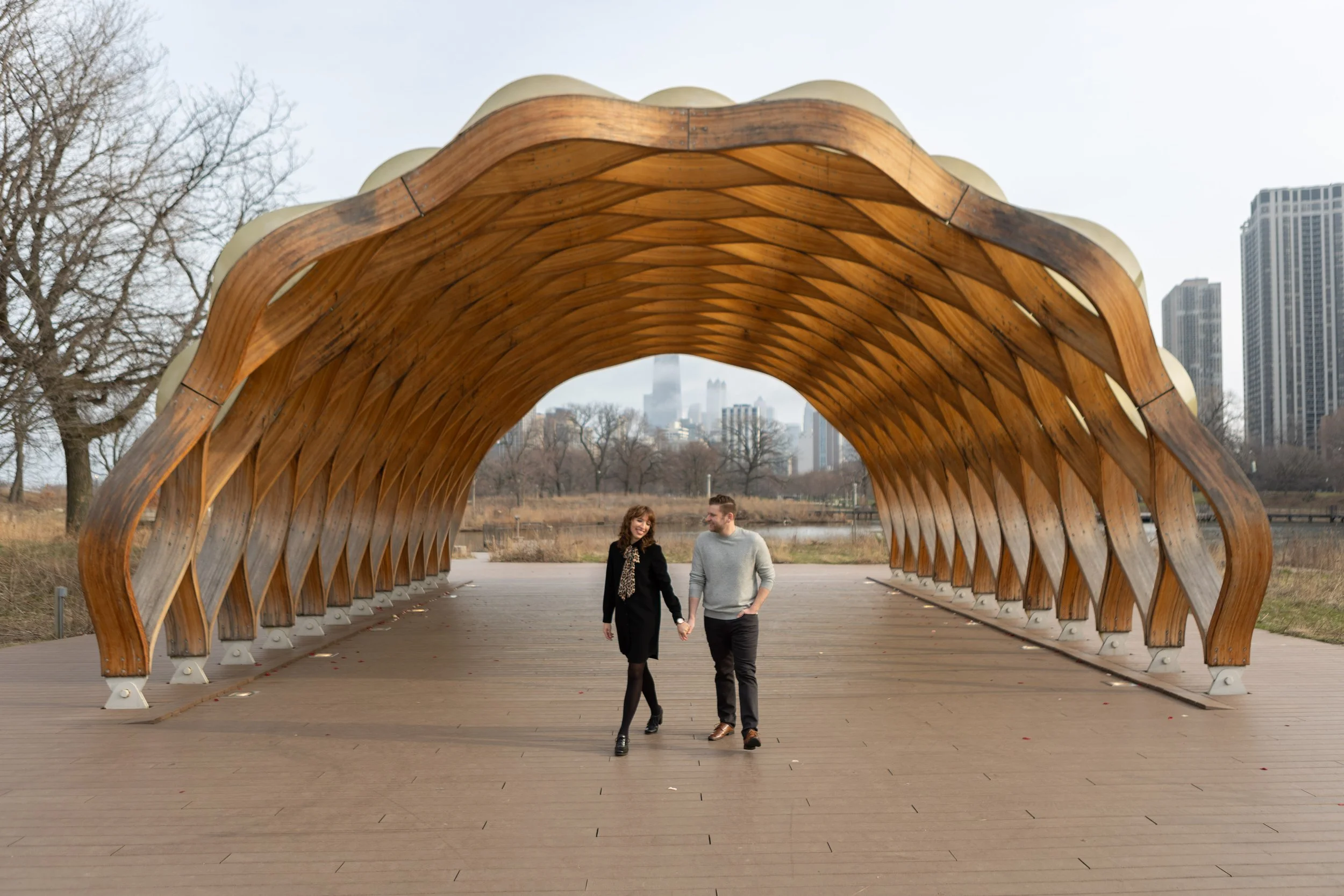 A couple walking hand in hand under a curved wooden pavilion in an urban park, with city skyscrapers in the background.