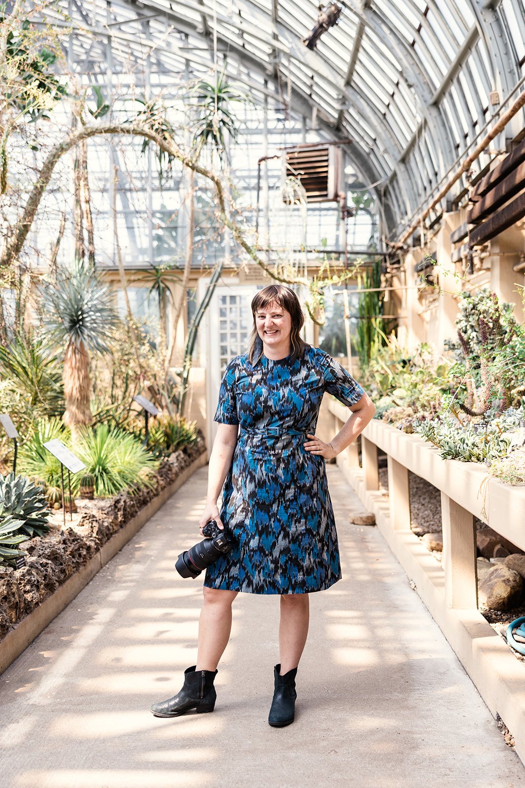 Alexis Ellers, a woman in a blue patterned dress and black ankle boots, standing inside the desert of the Garfield Park Conservatory, smiling and holding a camera.