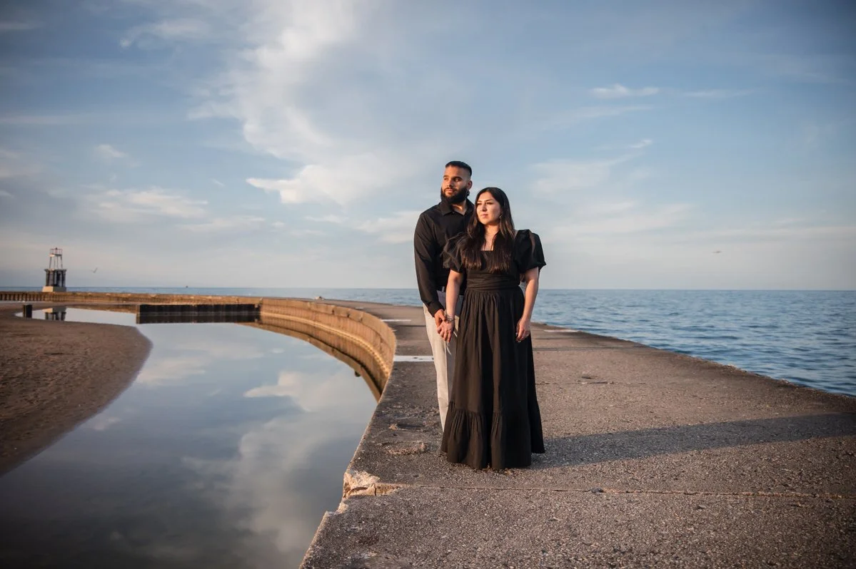 A man and woman both in black stand hand in hand on a concrete pier by Lake Michigan during sunset, with a lighthouse visible in the distance for their engagement photos.