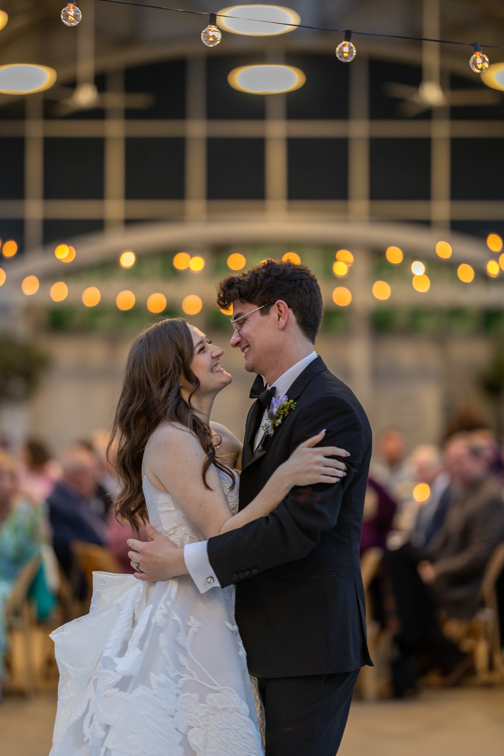 A bride and groom sharing their first dance at a wedding reception in a decorated, dimly lit venue with guests watching.
