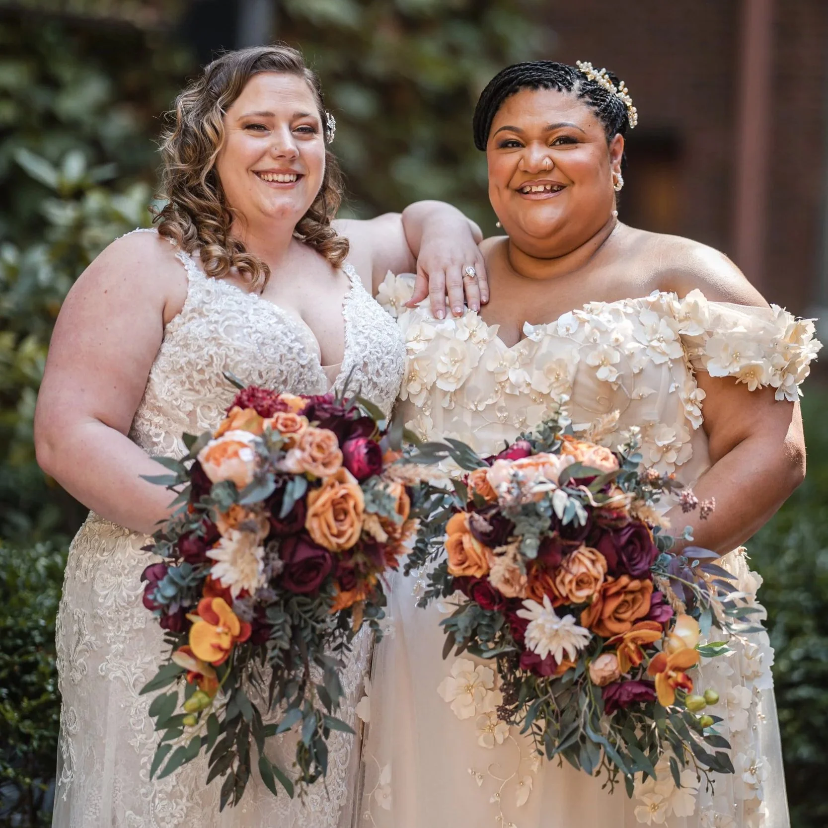 Two brides in wedding dresses holding floral bouquets, standing outdoors and smiling.