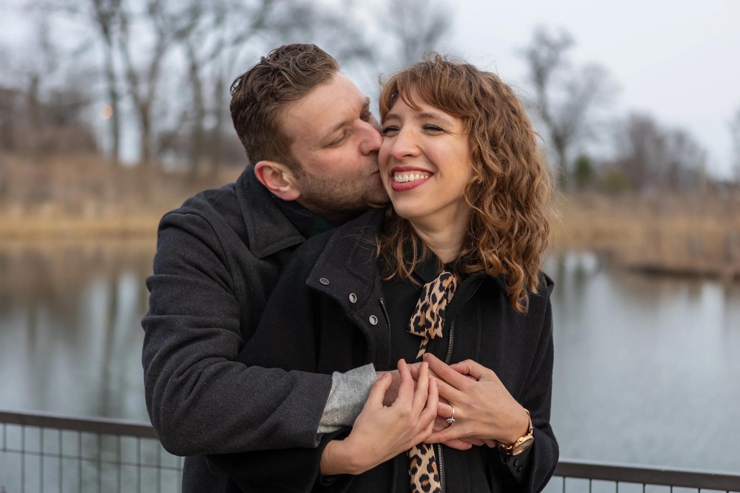 A couple standing by a river, with the man kissing the woman's cheek, both smiling and holding hands, in a chilly outdoor setting with trees and overcast sky.