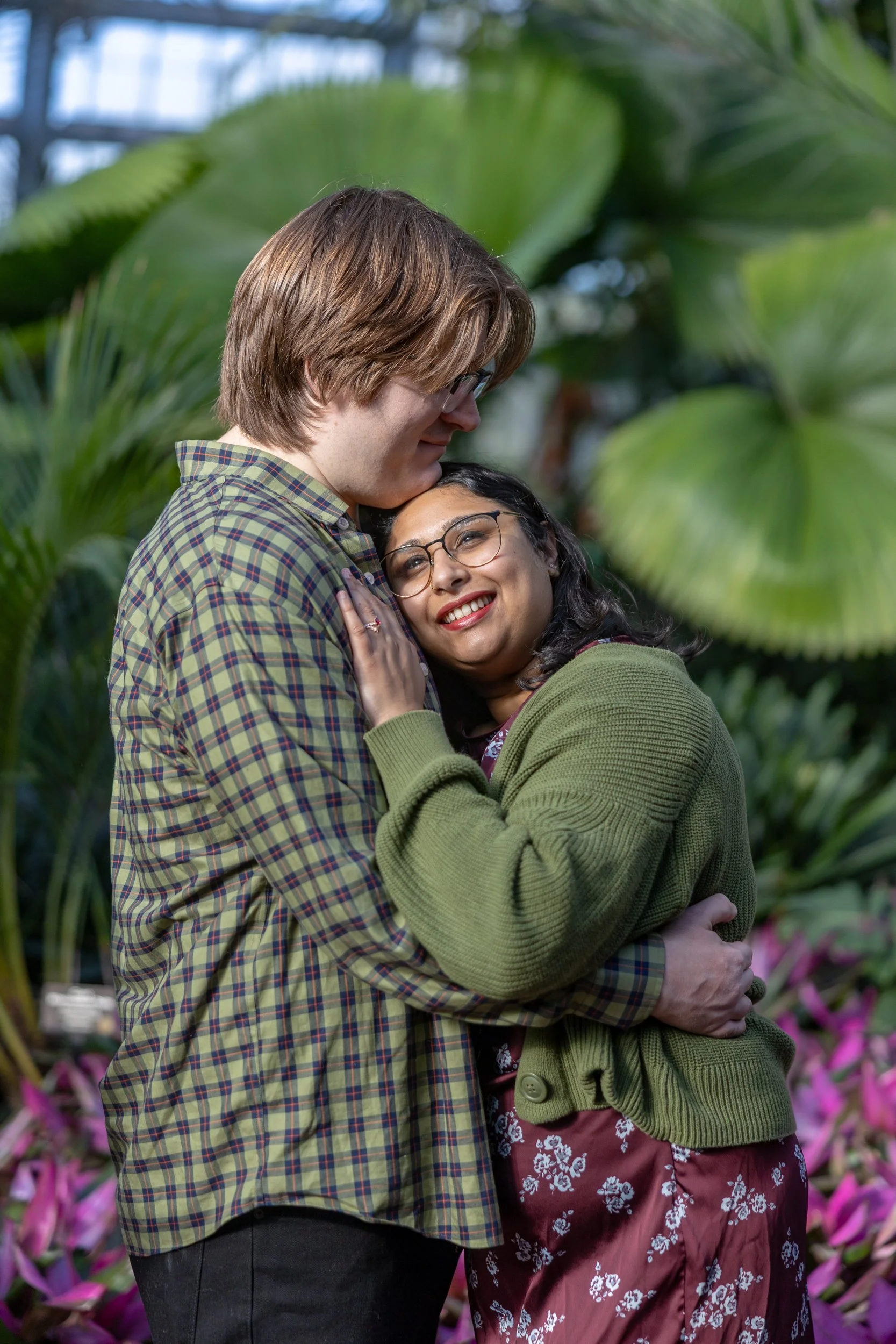 A couple hugging in a greenhouse or garden with lush green plants and pink flowers in the background, their faces close together, smiling and showing affection.