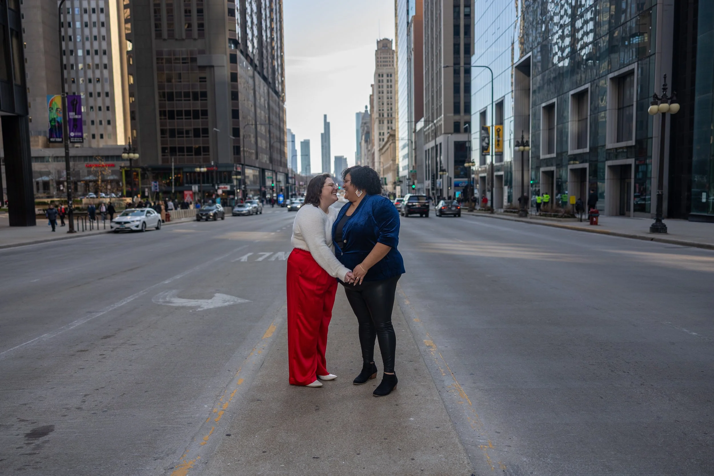 Two women holding hands and smiling at each other in the middle of a city street with tall buildings and cars in the background.