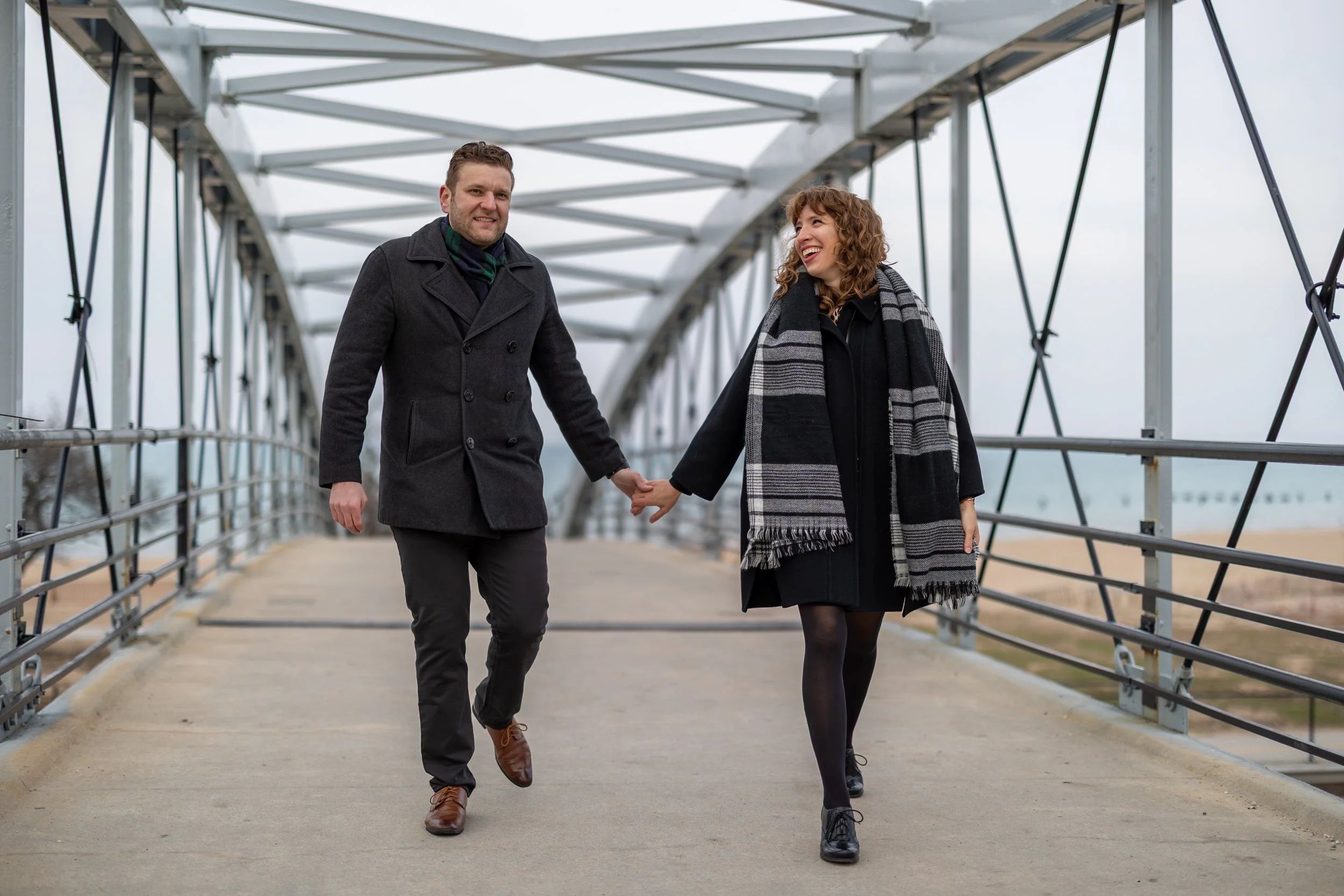 A couple holding hands and walking on a metal bridge, with the man on the left and the woman on the right, both smiling and dressed in warm, dark clothing.