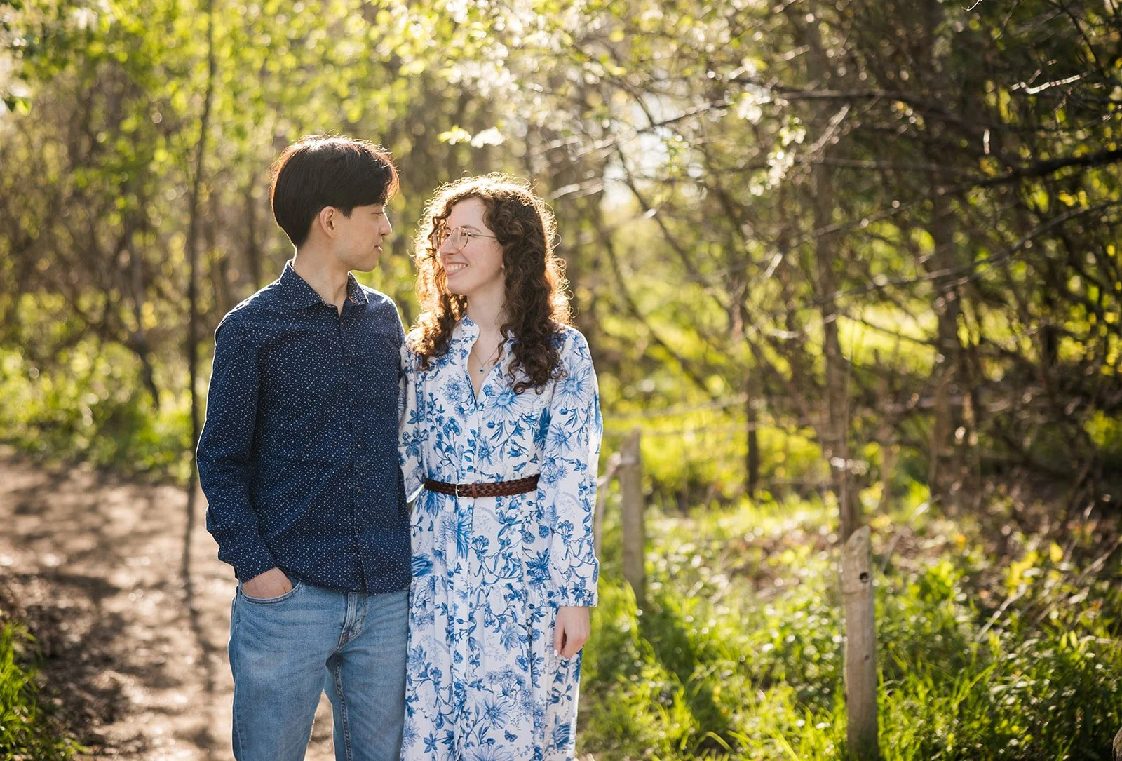 A young man and woman stand close together outdoors on a sunny day, looking at each other and smiling, surrounded by trees and greenery.