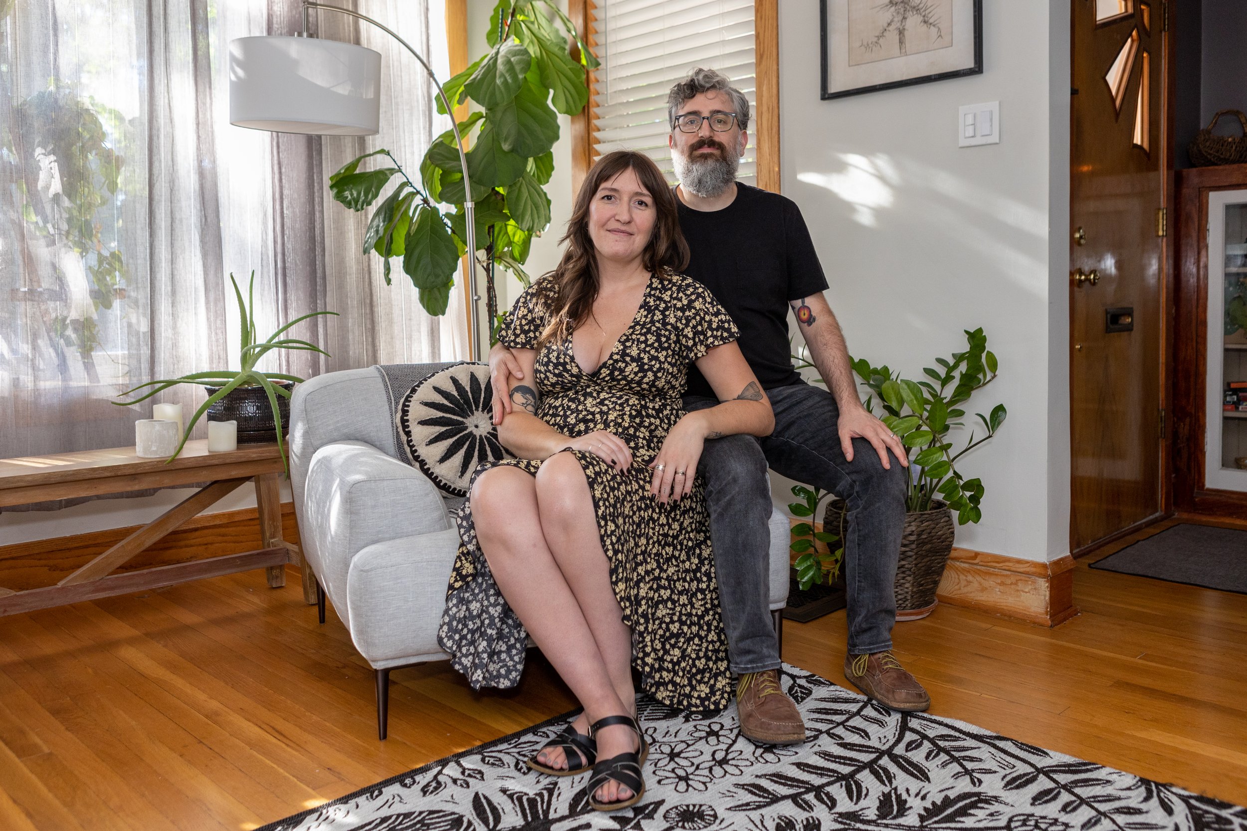 A man and woman sitting closely together on a couch in a well-lit living room with plants and wooden decor.