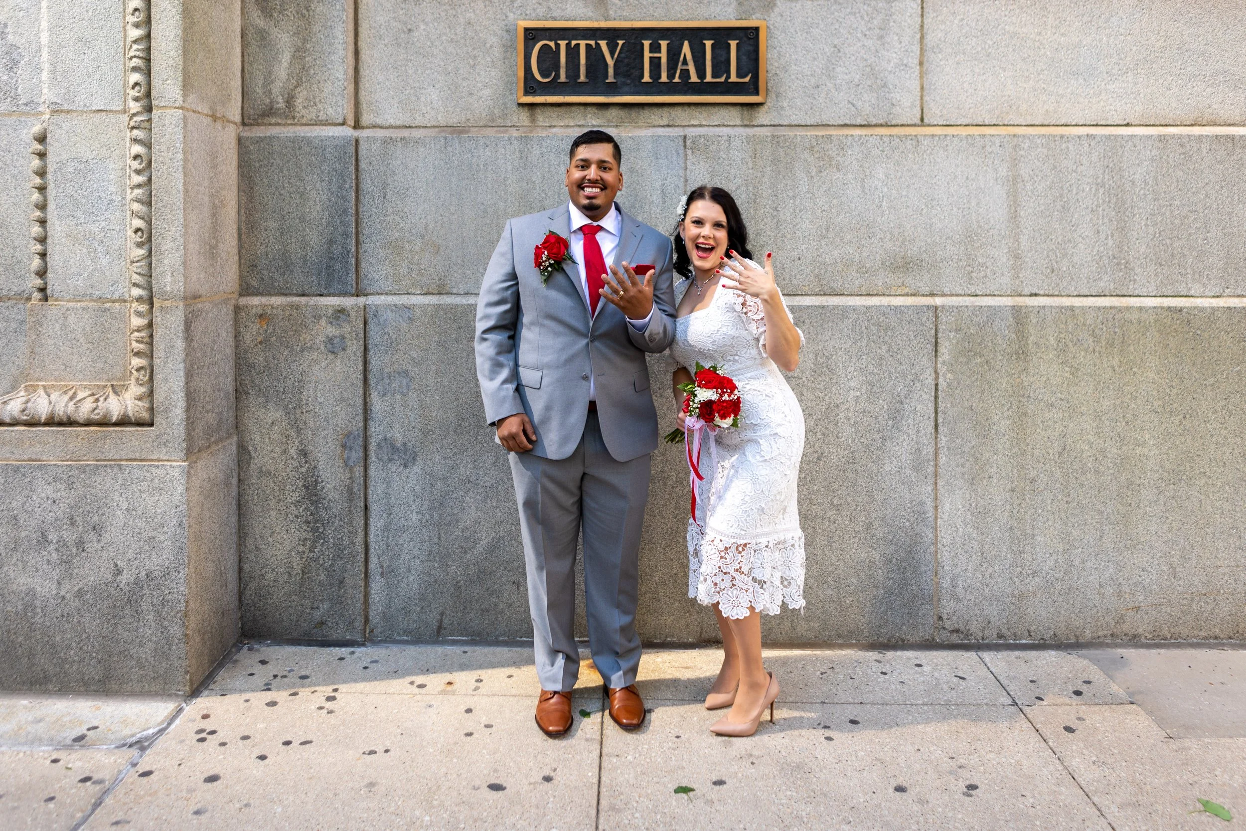 A happy couple dressed in wedding attire standing outside the City Hall, showing off their wedding rings and holding a bouquet of red and white flowers.