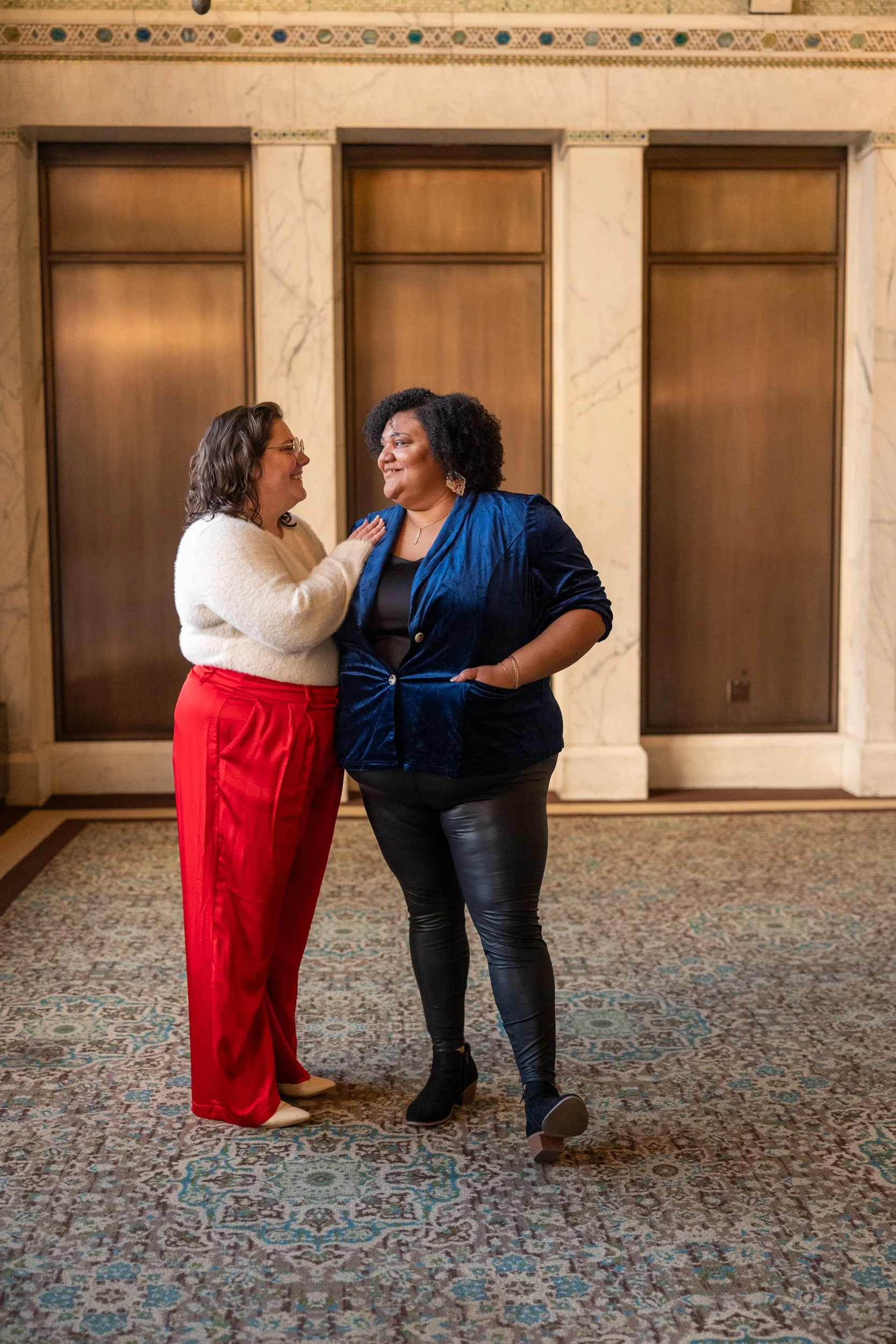 Two women standing and smiling at each other in an elevator lobby with marble walls and a patterned carpet.