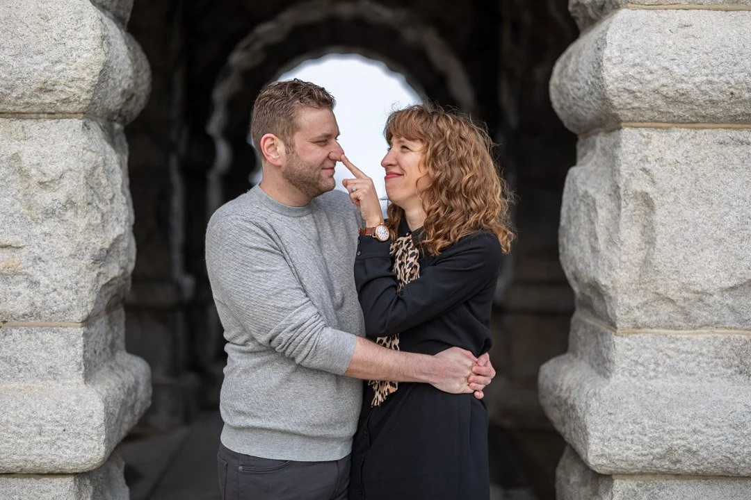 A couple standing close together through an archway, with the woman touching the man's nose with her finger, both smiling affectionately.
