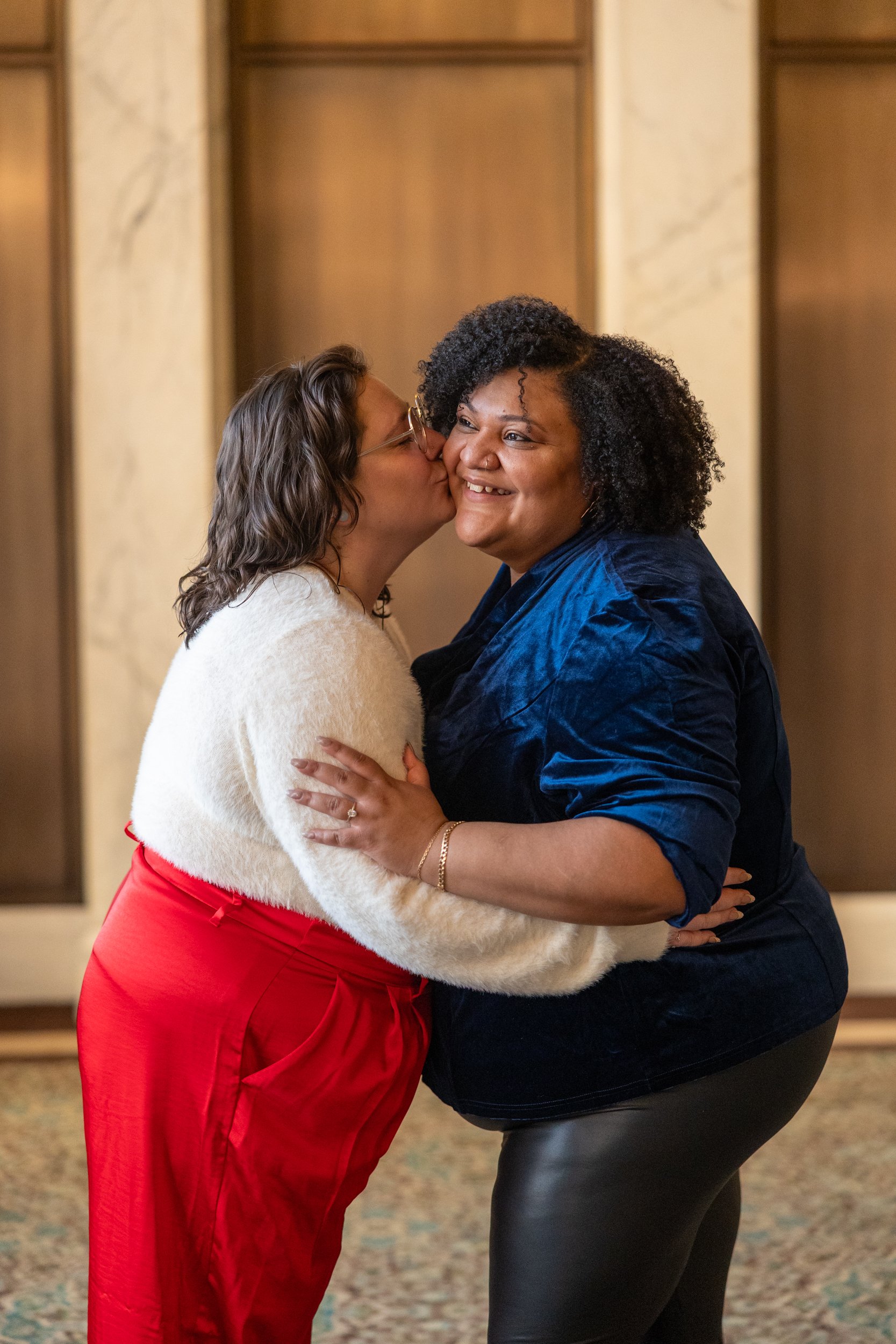 Two women sharing a warm embrace, one kissing the other's cheek, inside a warmly lit room with wooden paneling in the background.