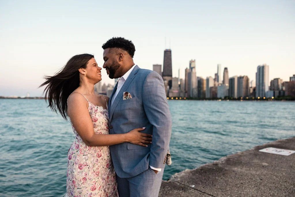 A couple standing close together near water with a city skyline in the background during sunset.
