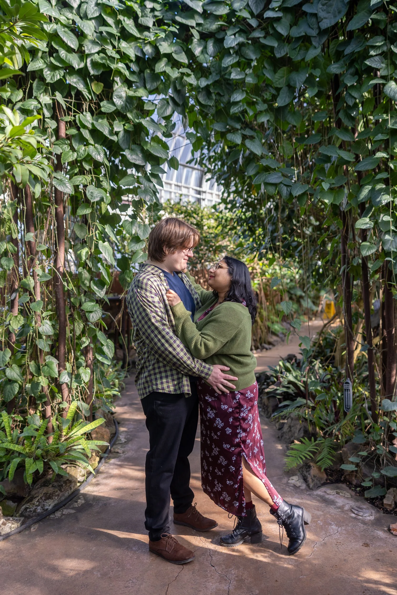 A couple standing close together in a lush greenhouse, looking into each other's eyes, surrounded by green plants and vines.