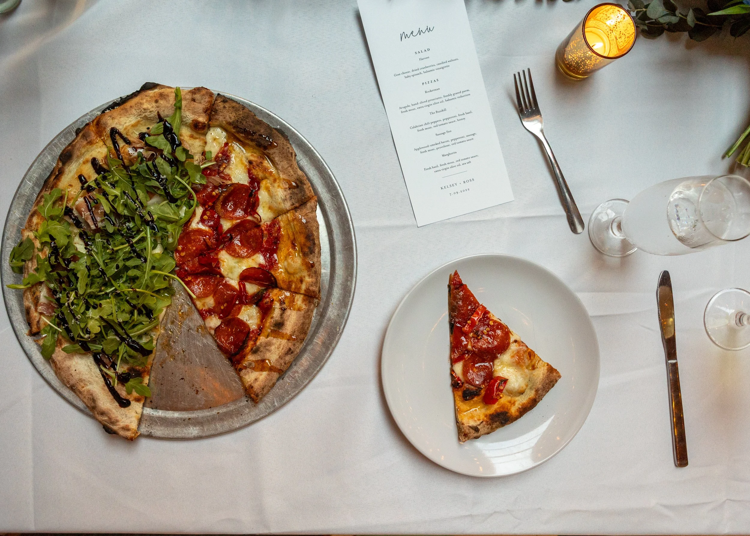A white tablecloth-covered table with a large pizza on a metal tray, a single slice of pizza on a white plate, a menu, a fork, a knife, a glass of water, a glass of sparkling wine, and a candle in a holder.