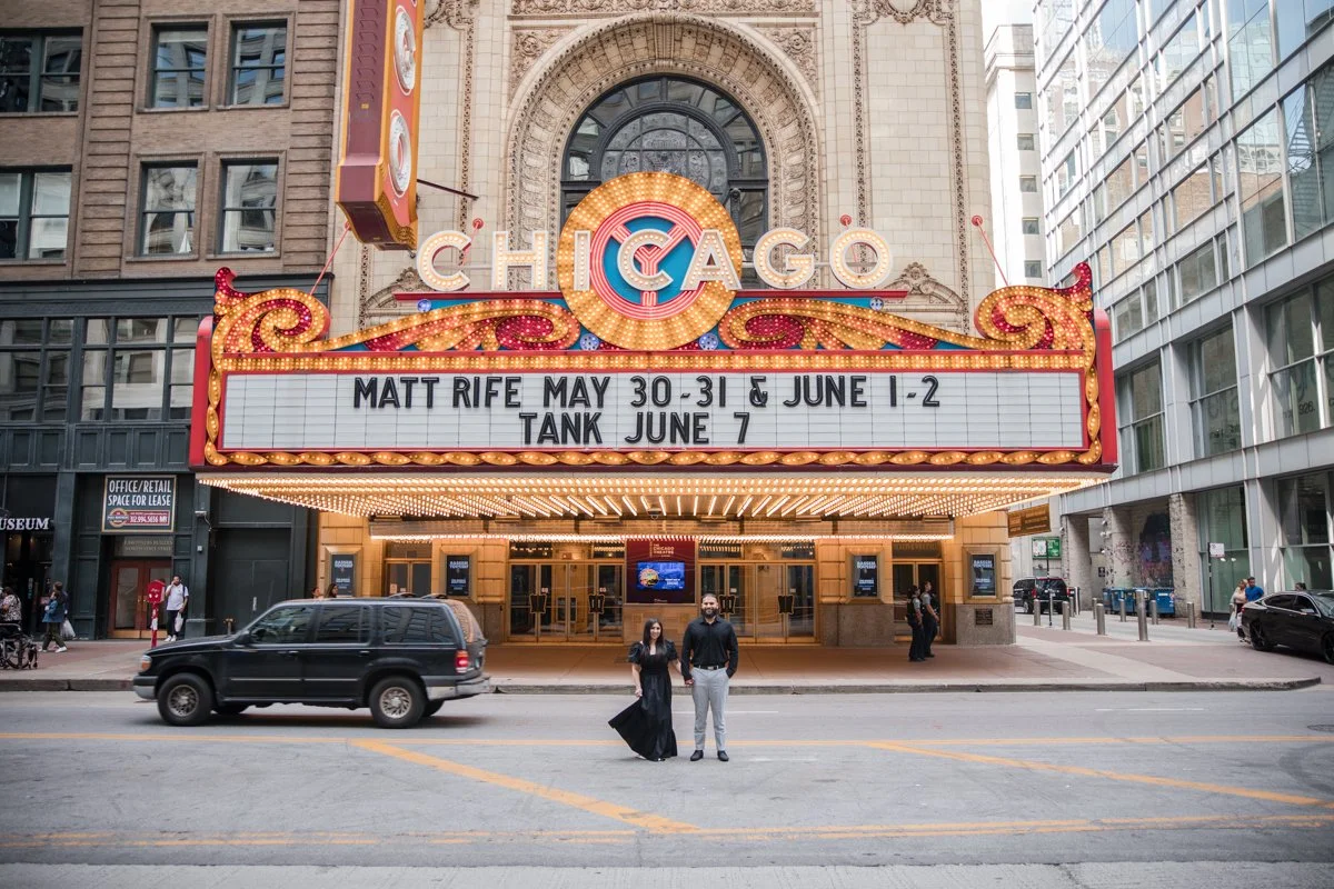 The Chicago theater marquee with upcoming show information and two people standing in the street in front, with a black SUV nearby.