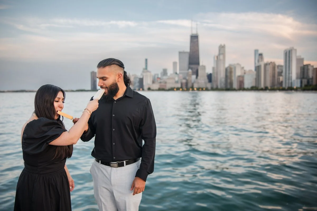 A woman is feeding ice cream to a man on a boat or near water with a city skyline, including the John Hancock Center, in the background.