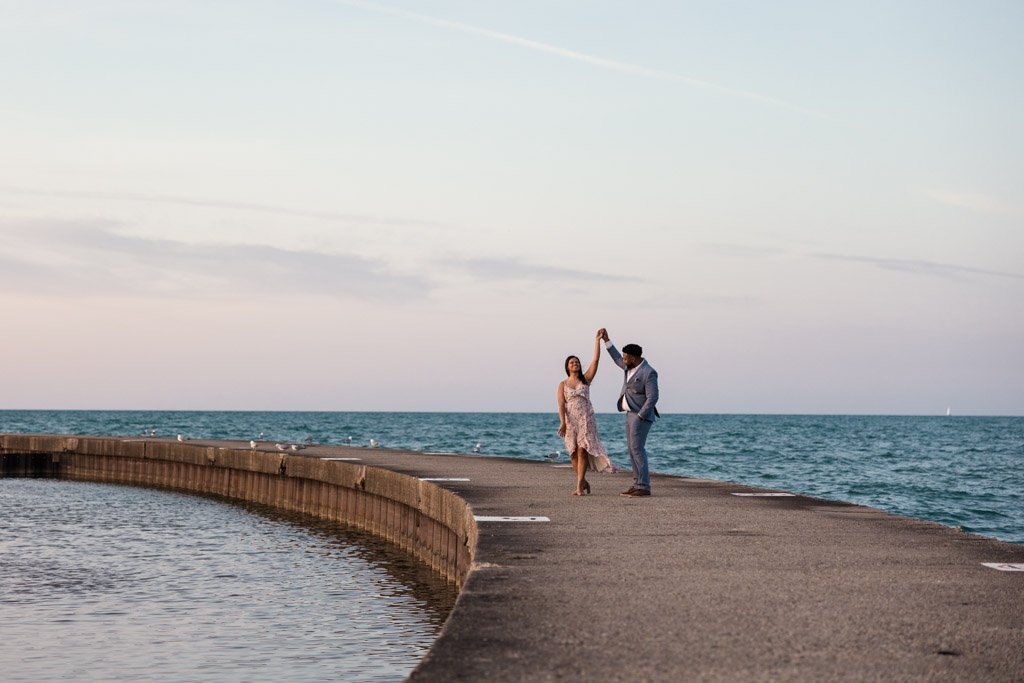 A couple dancing on a pier by the ocean during sunset, with the woman in a pink dress and the man in a suit and hat.