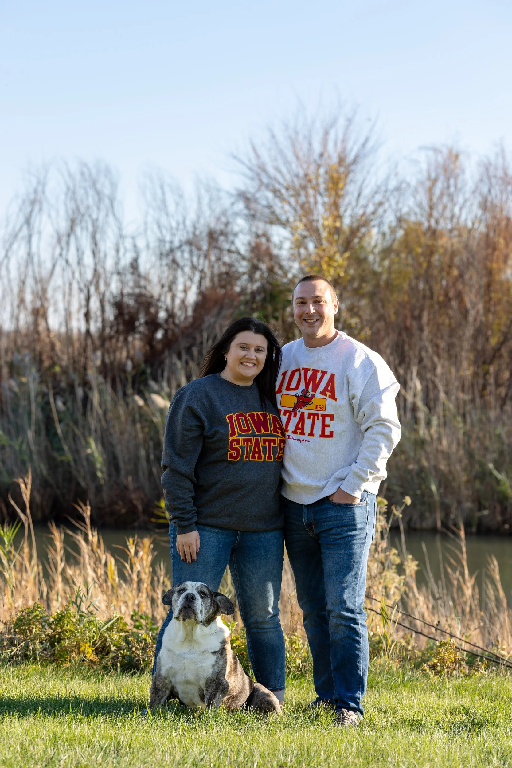 A couple standing outdoors on grass, wearing Iowa State sweatshirts, with a dog sitting in front of them, near a body of water and trees with autumn leaves in the background.