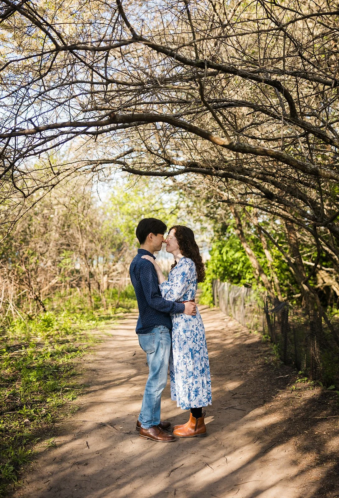 A couple standing close together on a dirt path under blooming trees, embracing lovingly in a park-like setting.
