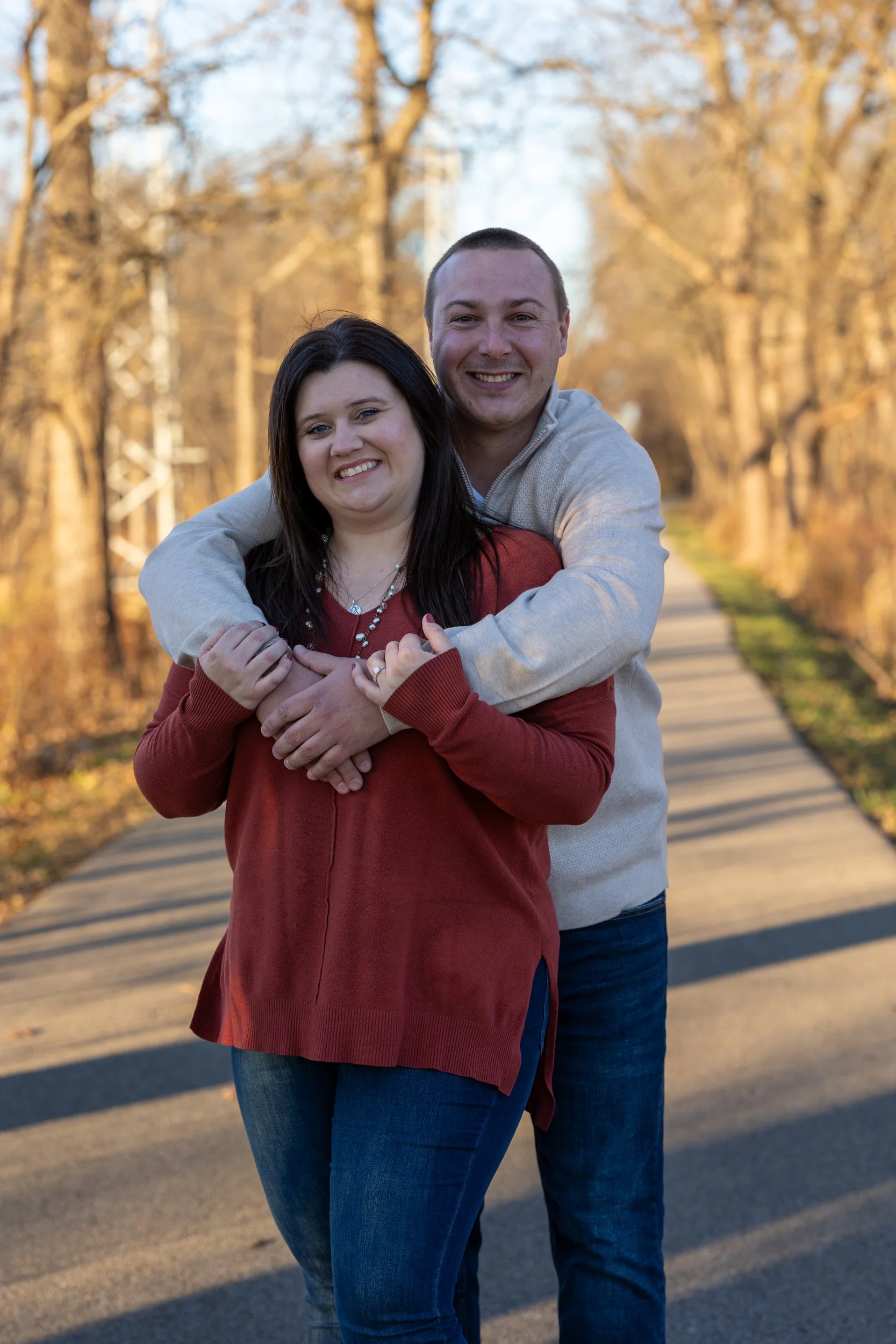 A happy couple standing on a tree-lined sidewalk in autumn, with the man hugging the woman from behind.