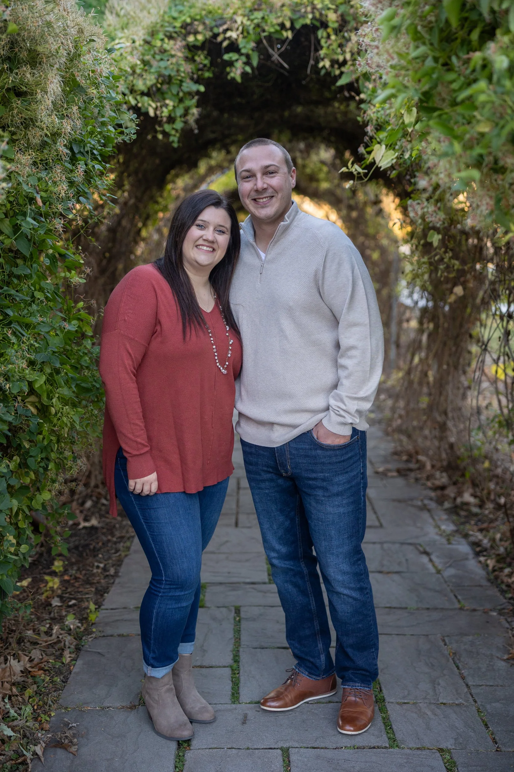 A smiling couple standing together on a stone pathway covered by a natural arch of greenery and flowers.
