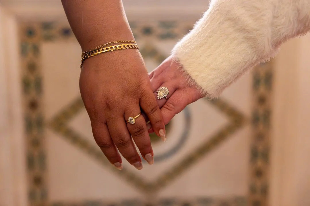 Two hands with rings and jewelry gently touching, one hand with darker skin tone and the other with lighter skin tone, against a blurred patterned background.