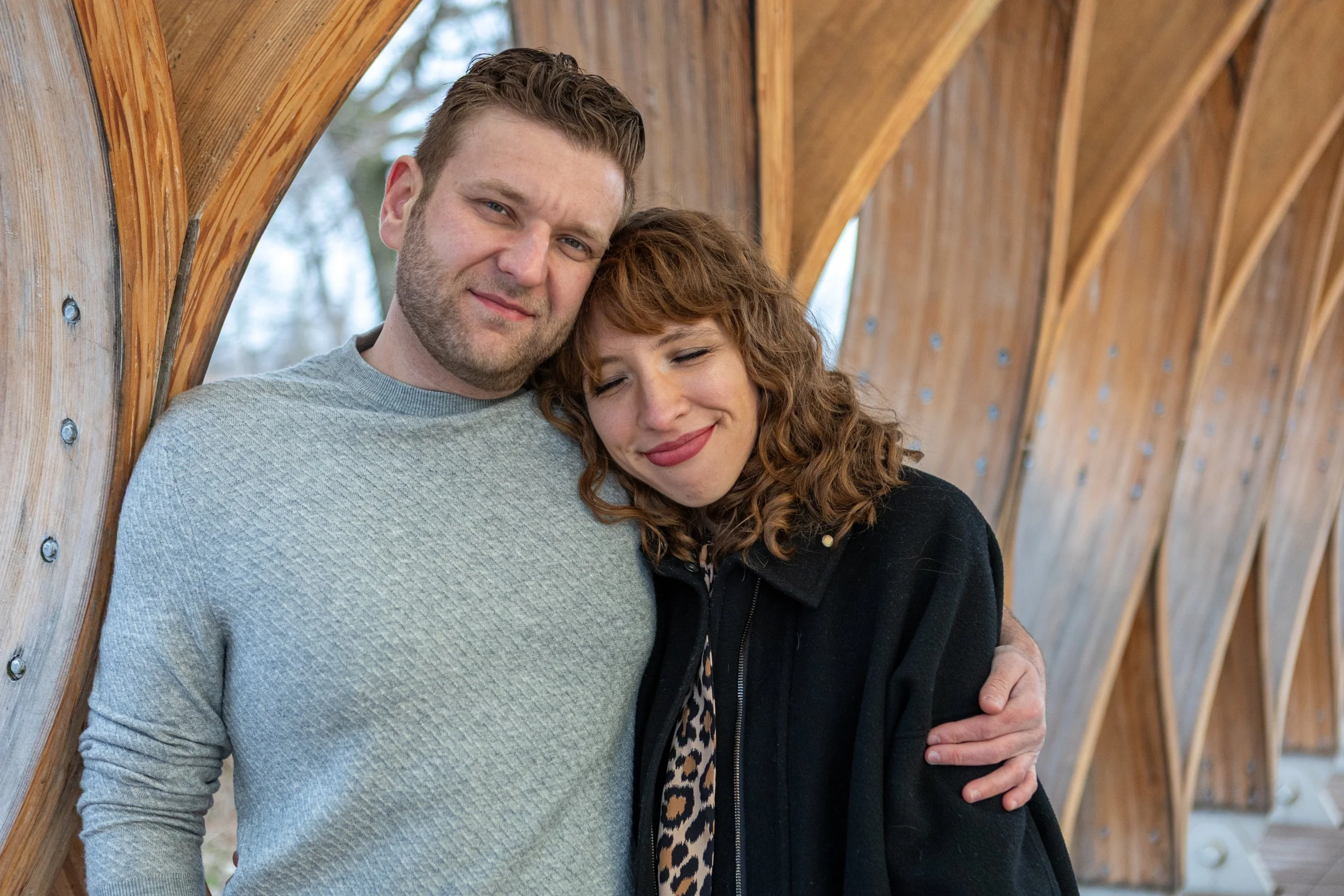 A man and woman sharing a hug, smiling, standing under a wooden archway.