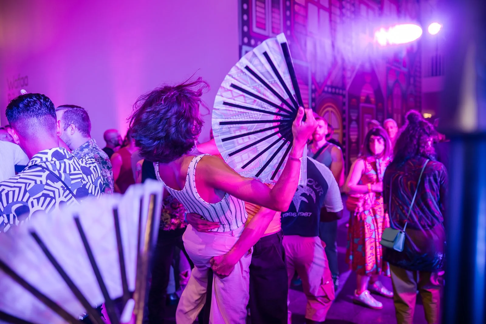 Queer people dancing at a party in the Museum of Contemporary Art with purple lighting, some holding folding fans, in a colorful, vibrant setting.